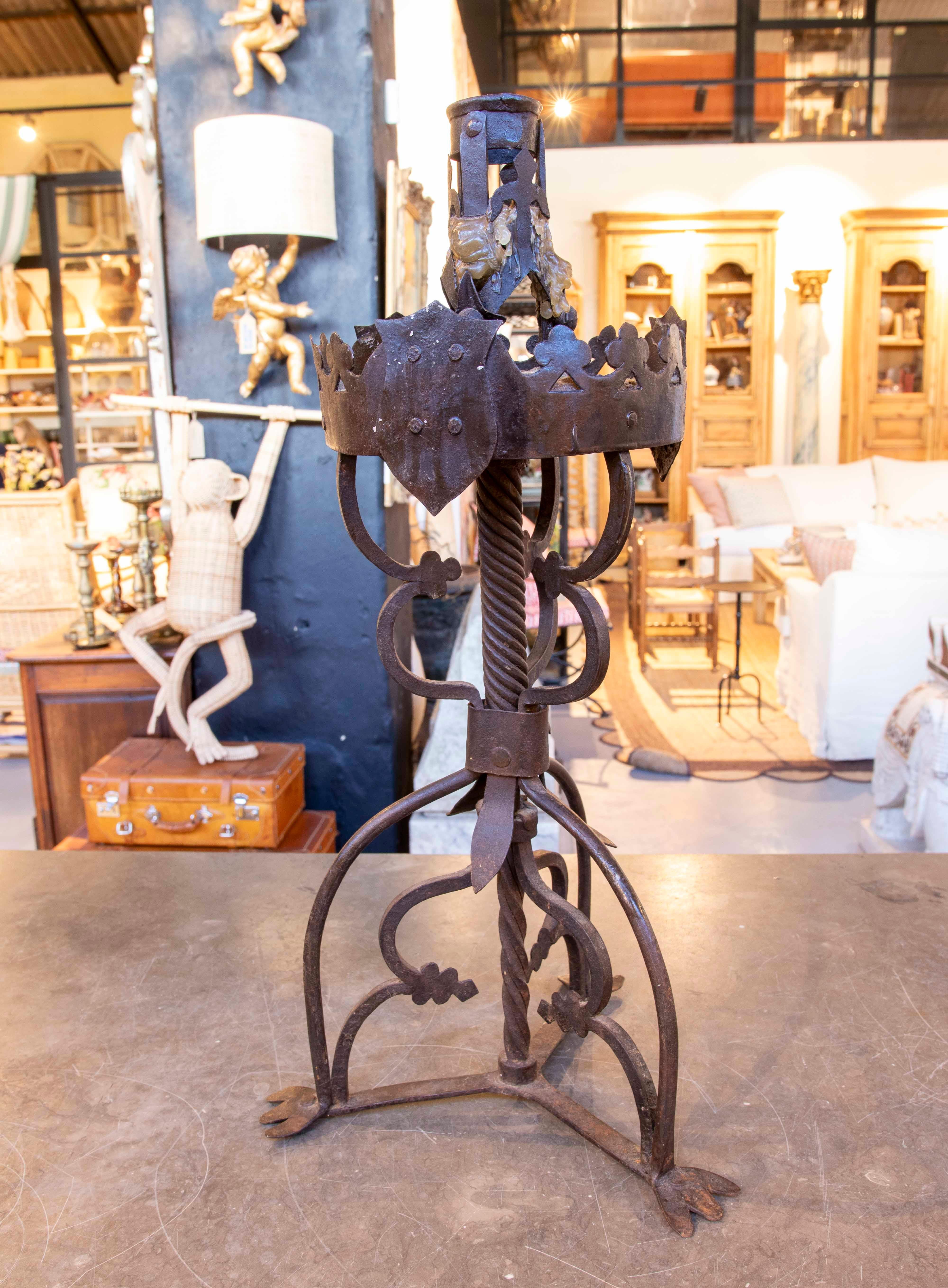 Un imponente candeliere in ferro battuto lavorato a mano, con un design robusto e dettagli ornamentali che ricordano lo stile medievale. La struttura presenta una base decorata a tre gambe, un'asta centrale contorta ed elementi decorativi a forma di
