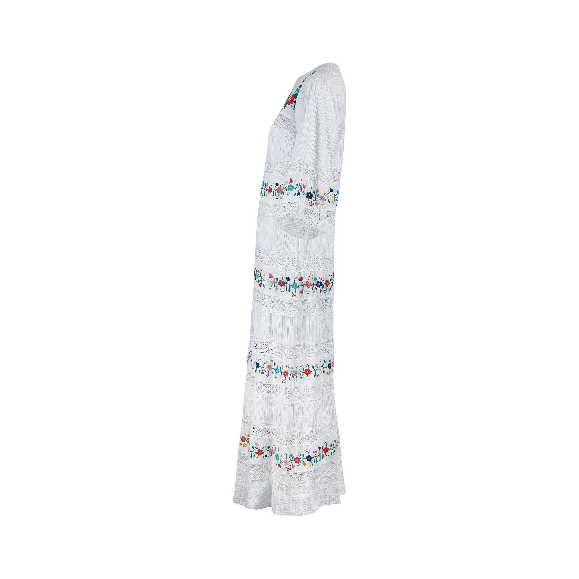 Dieses knöchellange Hochzeitskleid von Yolanda aus den 1970er Jahren stammt aus Mexiko und weist viele Merkmale des traditionellen mexikanischen Designs auf. Es ist in einer luftigen A-Linien-Silhouette aus weißer Baumwolle geschneidert und über und