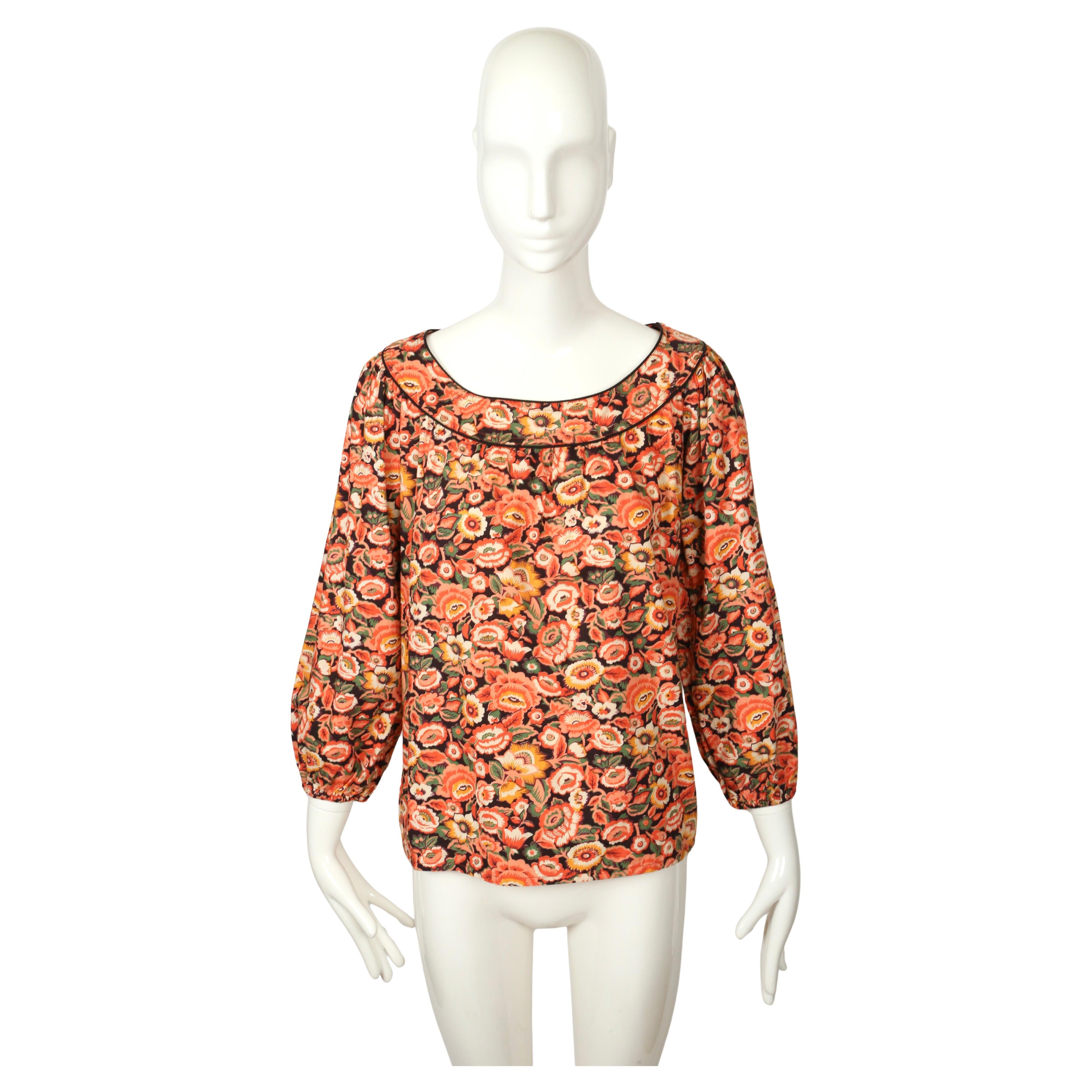 Blusa de algodón SAINT LAURENT de los años 70 con un alegre estampado floral en tonos cálidos de coral, naranja, amarillo y verde sobre fondo negro. El diseño presenta una silueta relajada con un amplio escote barco perfilado con un fino ribete