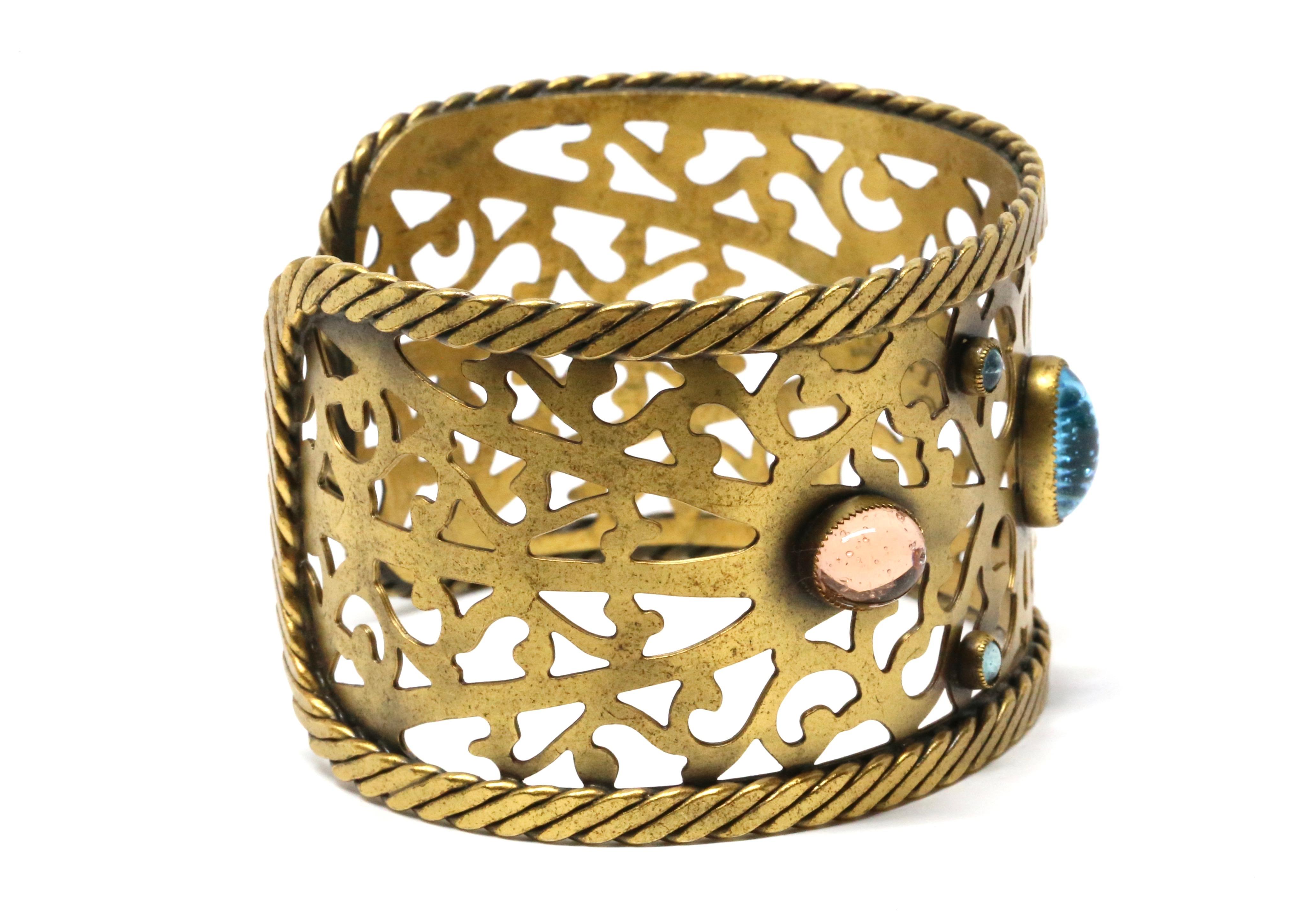Yves Saint Laurent, 1970's

An ornate openwork cuff bracelet from Yves Saint Laurent, crafted in gilt metal with an intricate pierced design. The wide band is bordered by twisted rope edging and embellished with colorful poured glass cabochons,