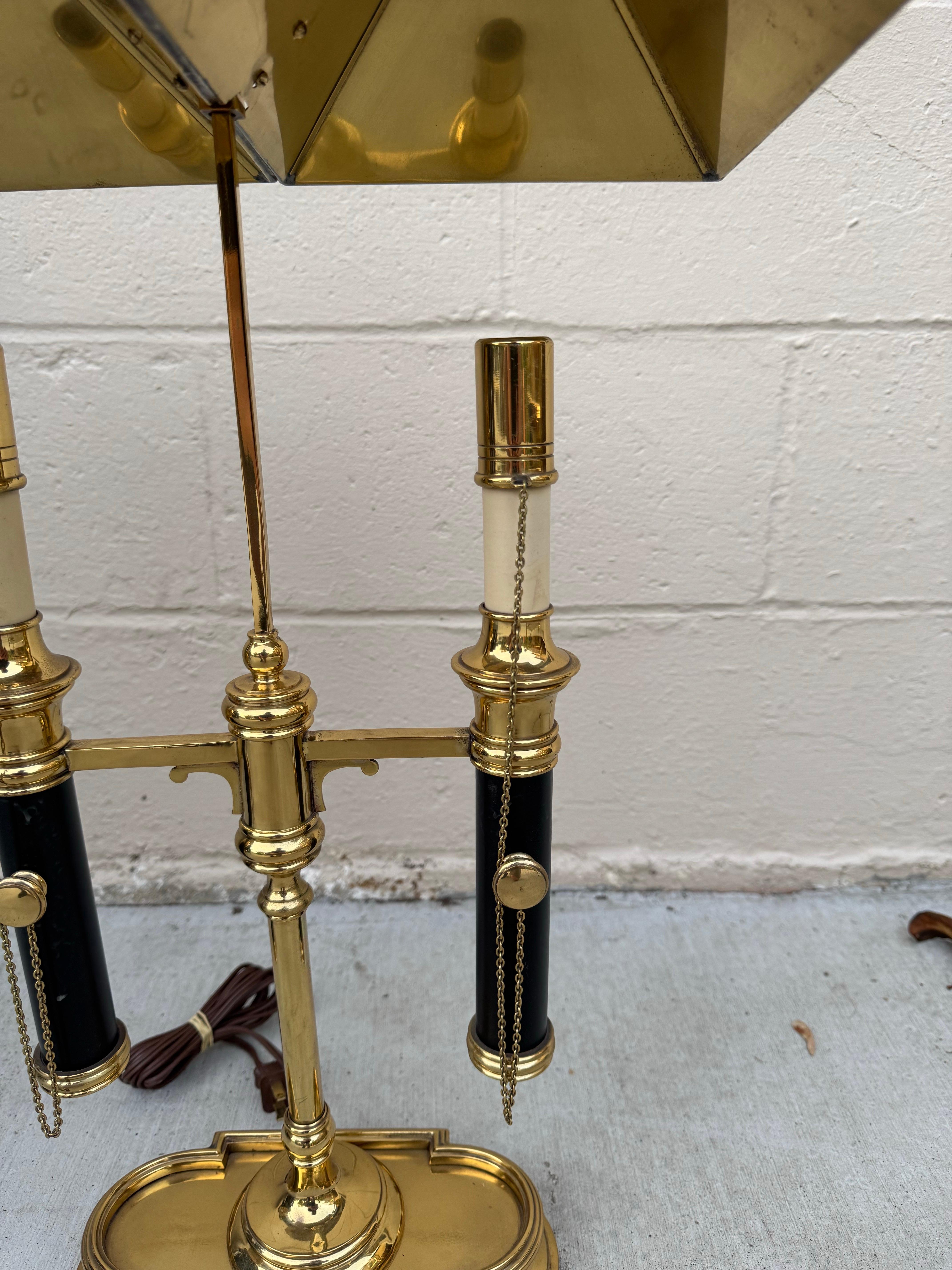 Impresionante lámpara de sobremesa estilo bouillotte Chapman de 1972, con una elegante estructura de latón y detalles clásicos de esmalte negro. La lámpara presenta dos columnas ajustables en forma de vela, cada una con cadenas, bajo una doble