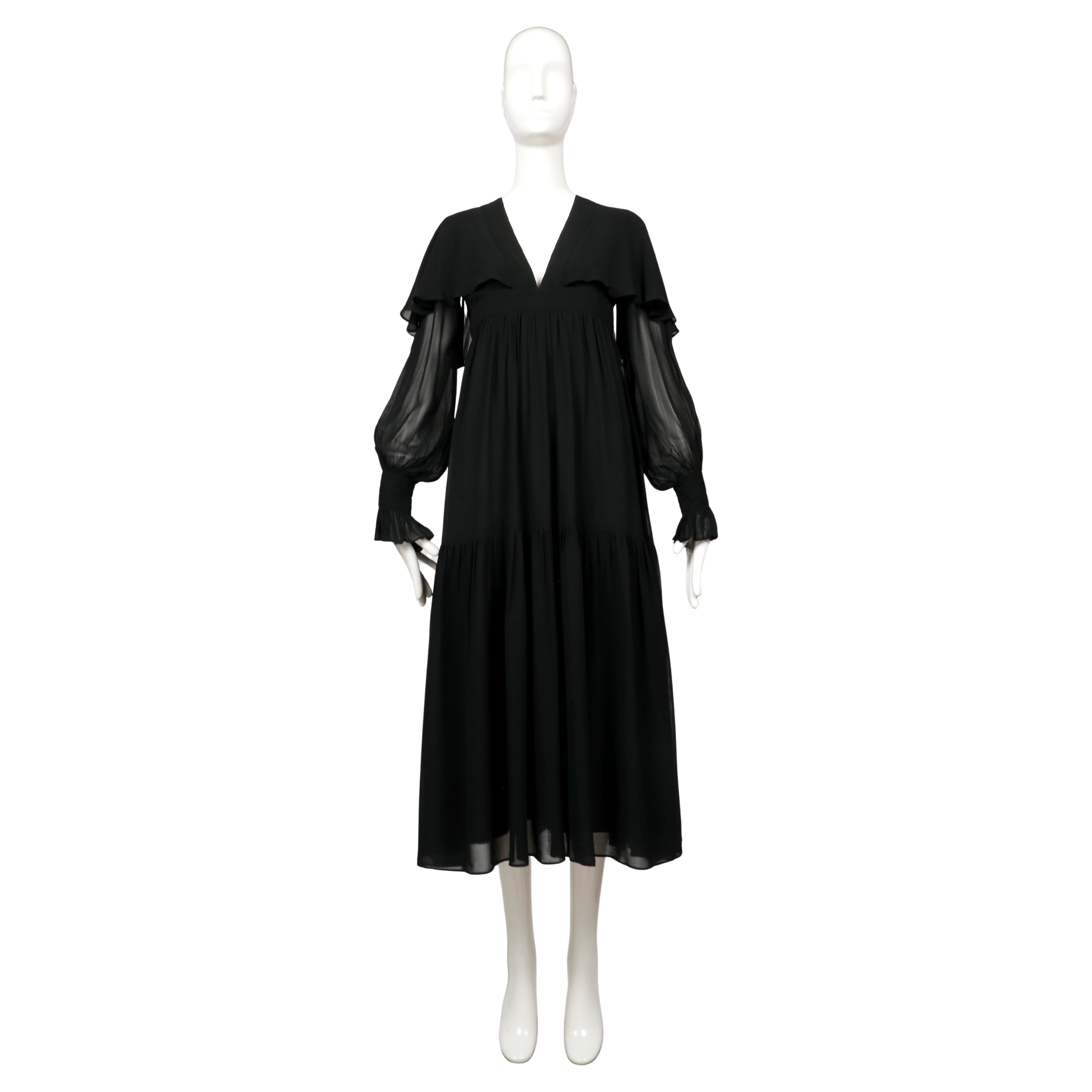 Superbe robe de soirée en mousseline de soie noire d'Ossie Clark datant de 1972. Conçue avec une taille empire, une jupe fluide à étages, des manches blouson transparentes terminées par un élastique aux poignets et une cape douce qui se drape sur