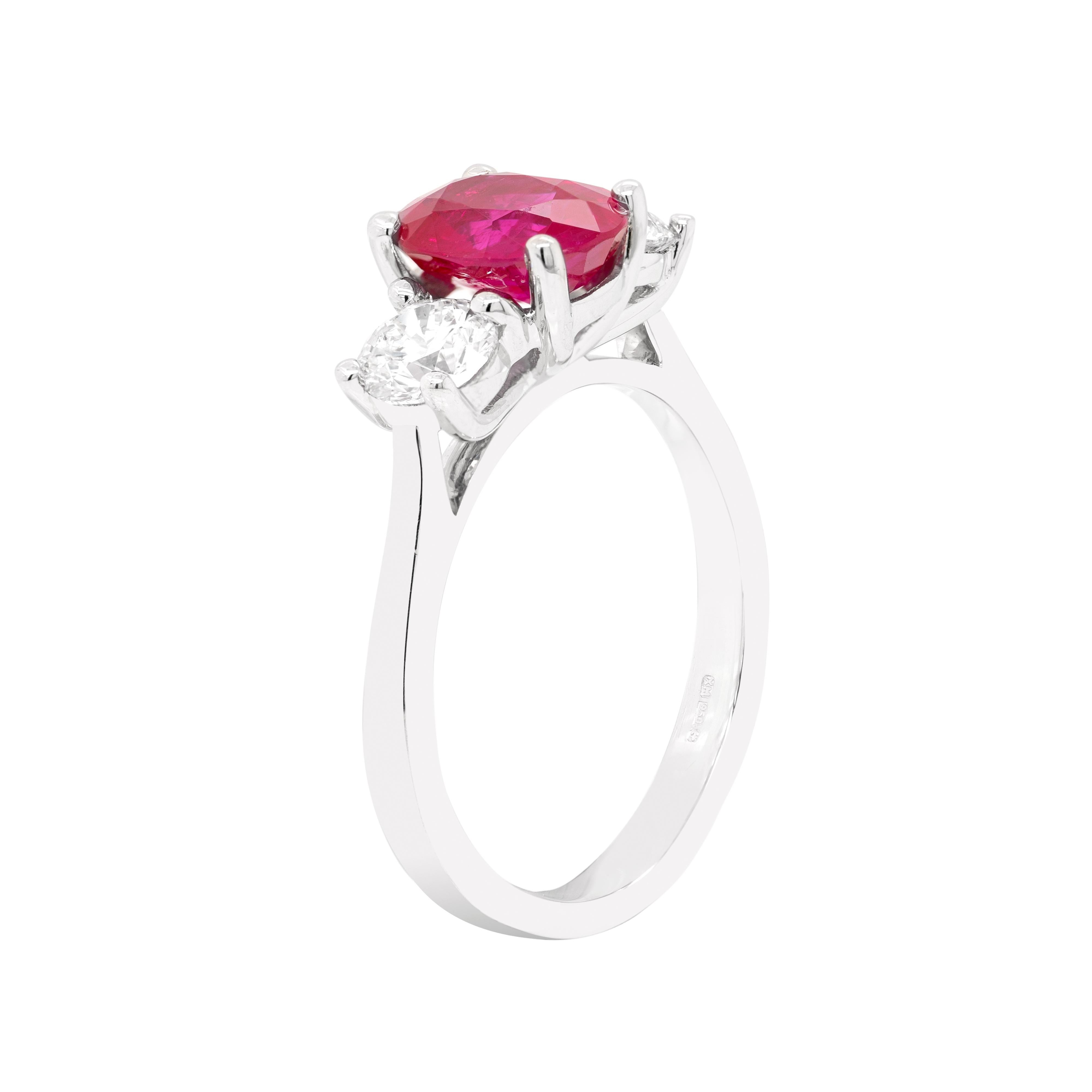 Exquisite platinum engagement ring featuring a 1.97ct vibrant oval shaped ruby in the centre, in a four claw, open back setting. The beautiful ruby is accompanied by two round brilliant cut diamonds, one on either side, with a total combined weight