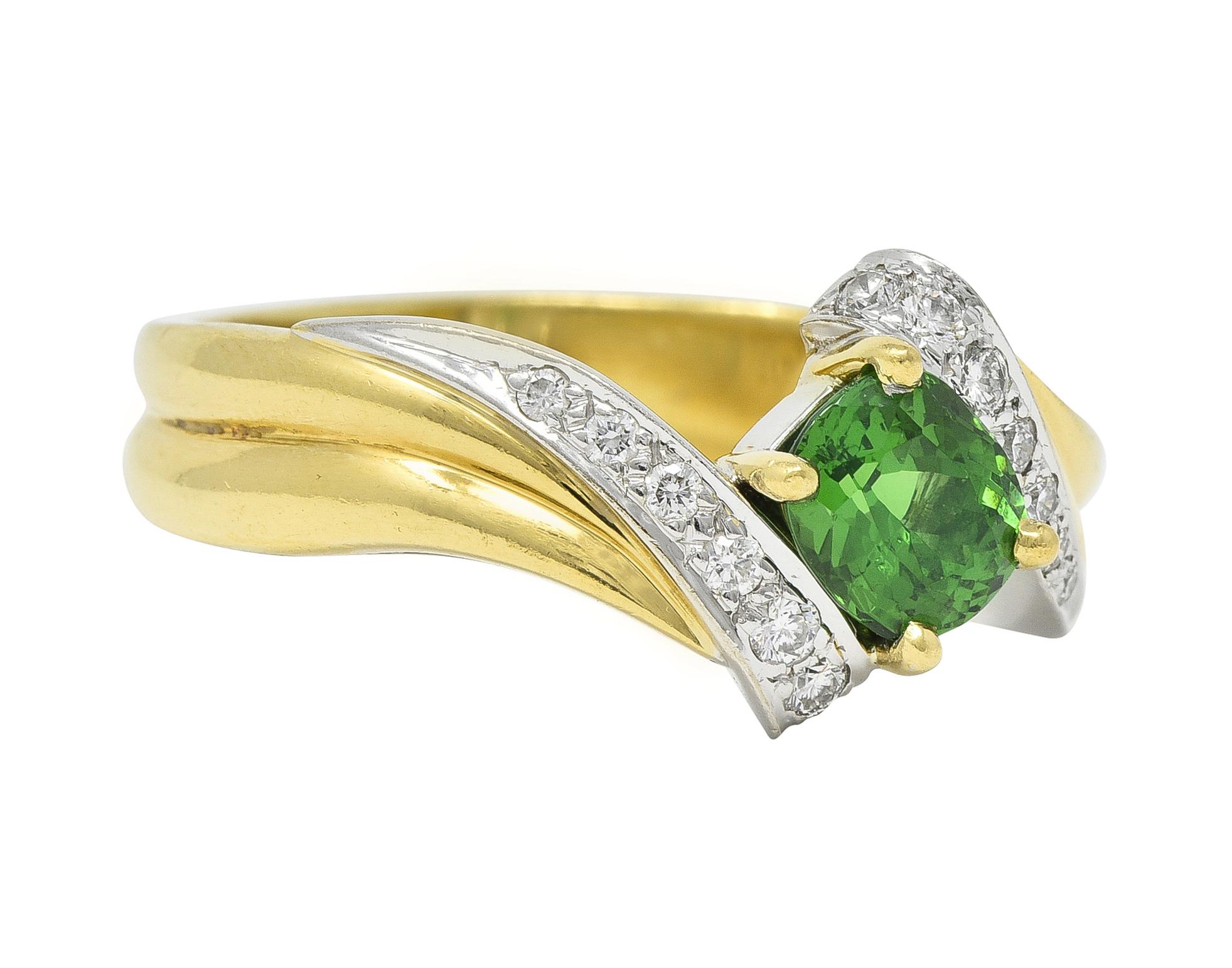 Centering a cushion cut tsavorite garnet weighing approximately 1.40 carats 
Transparent bright green in color - prong set in basket
Flanked by swirling bypass style shoulders 
Grooved with white gold segments
Bead set with round brilliant cut