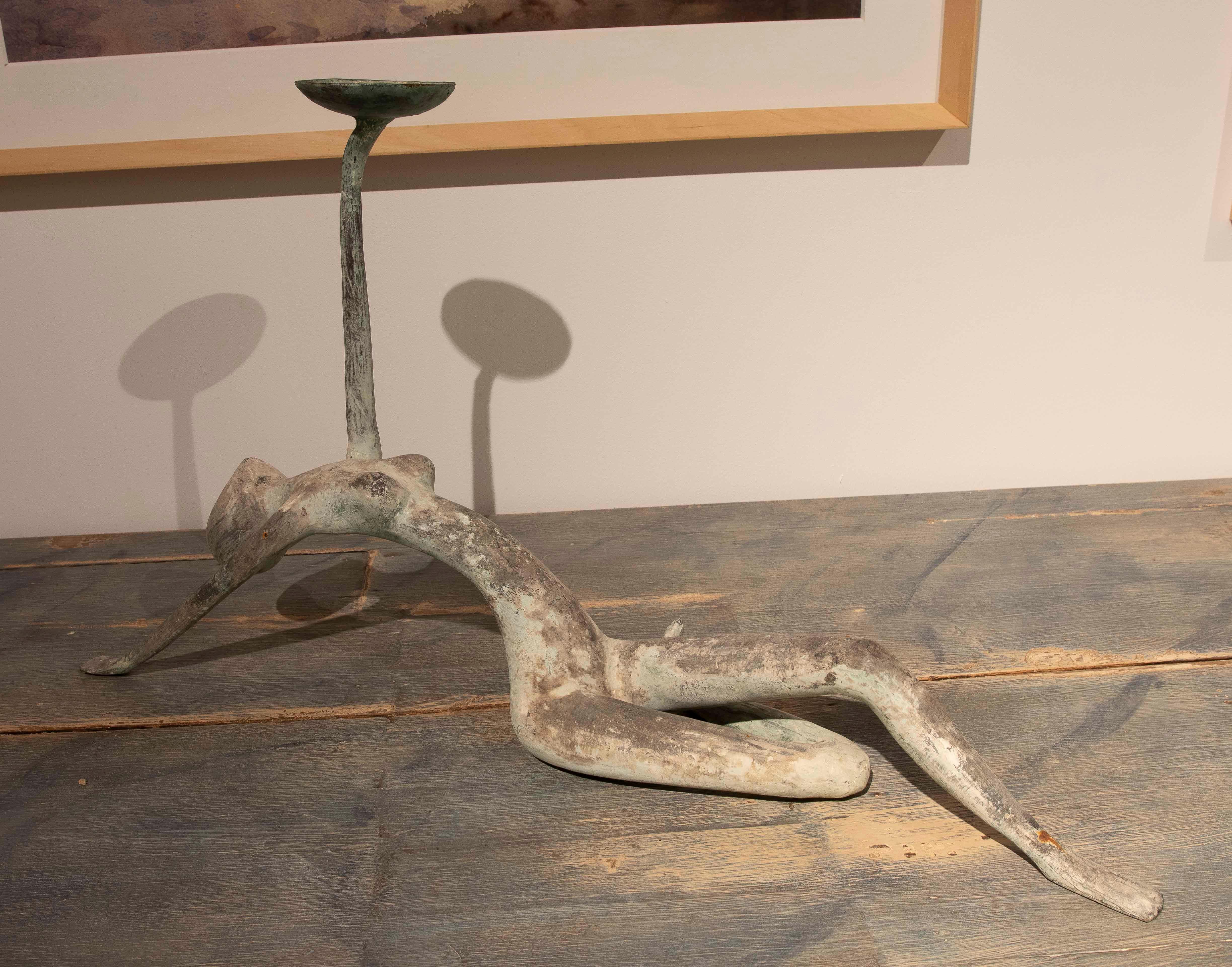 This intriguing bronze sculpture depicts a stylized human figure in a fully extended pose, supporting a circular base on an elevated leg. Its abstract design, combined with an aged finish revealing greenish hues, evokes a sense of movement and