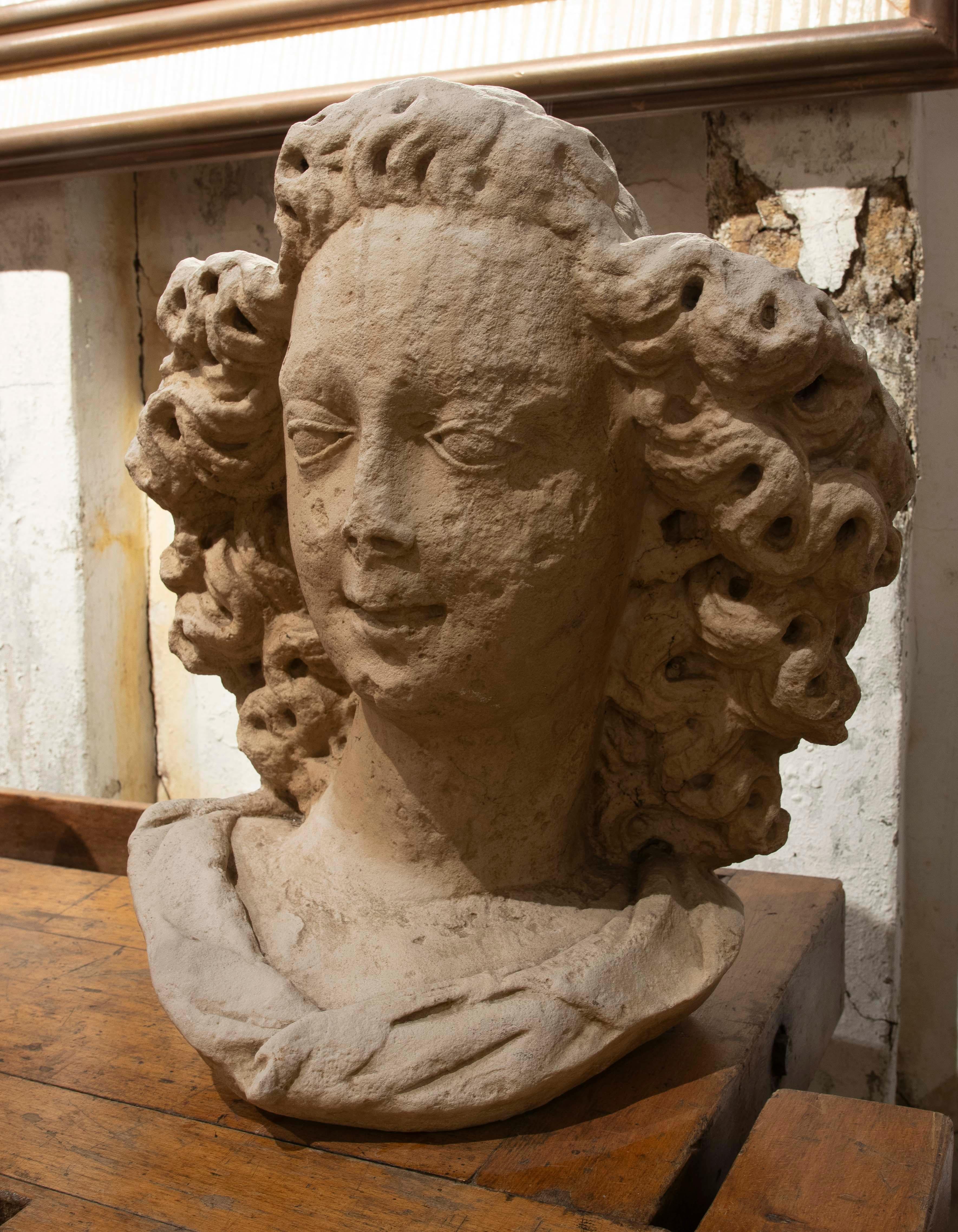 Scultura in pietra raffigurante il busto di un angelo con un'espressione serena e capelli ricci intricatamente scolpiti. Il viso presenta tratti morbidi e uno sguardo introspettivo, mentre il busto mostra un panneggio classico con pieghe