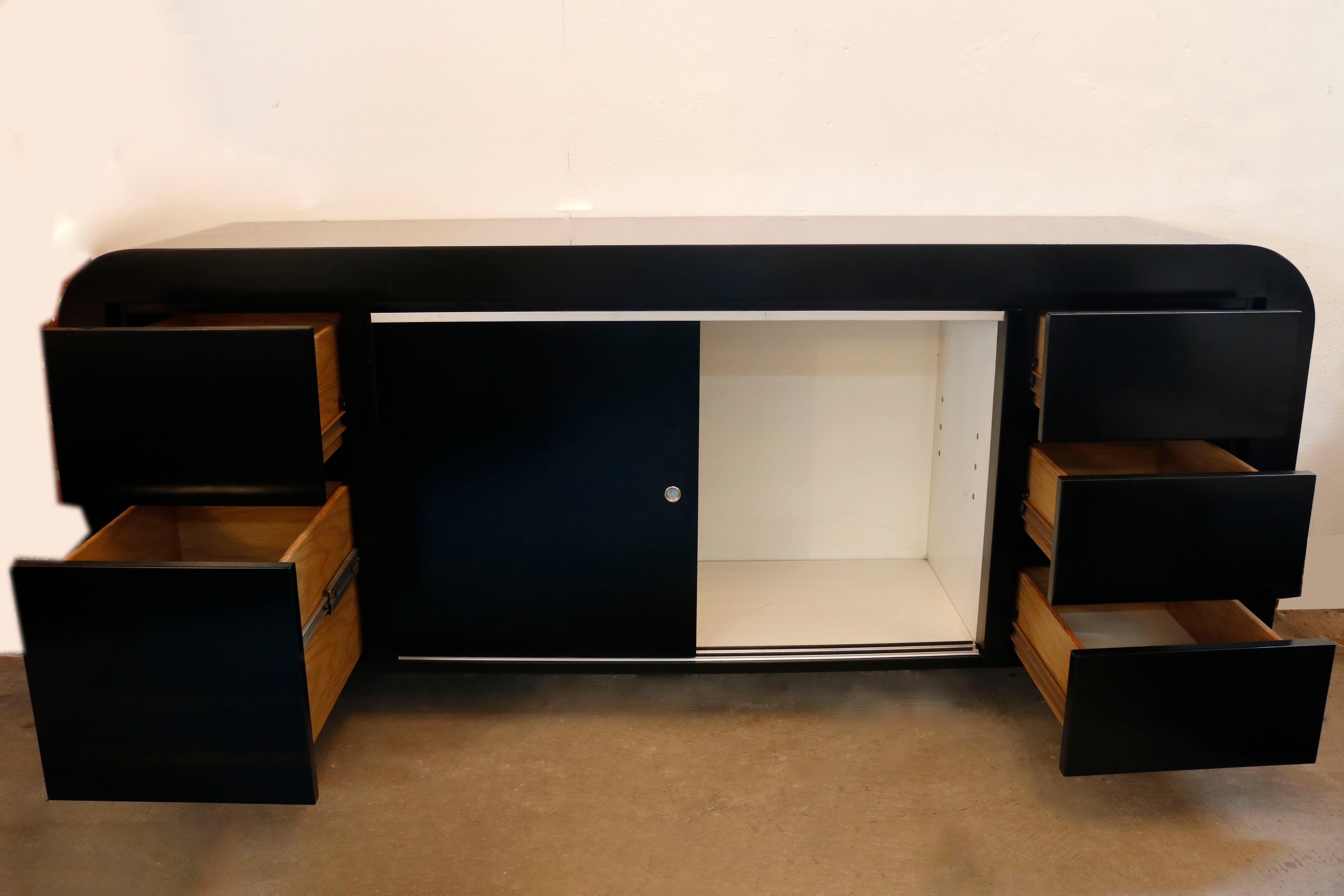This lustrous, black lacquered credenza, complete with smooth round edges, has ample storage space including 3 drawers on the right side and 2 file drawers on the left. The middle cabinet features sliding doors opening on a shelf. The piece has