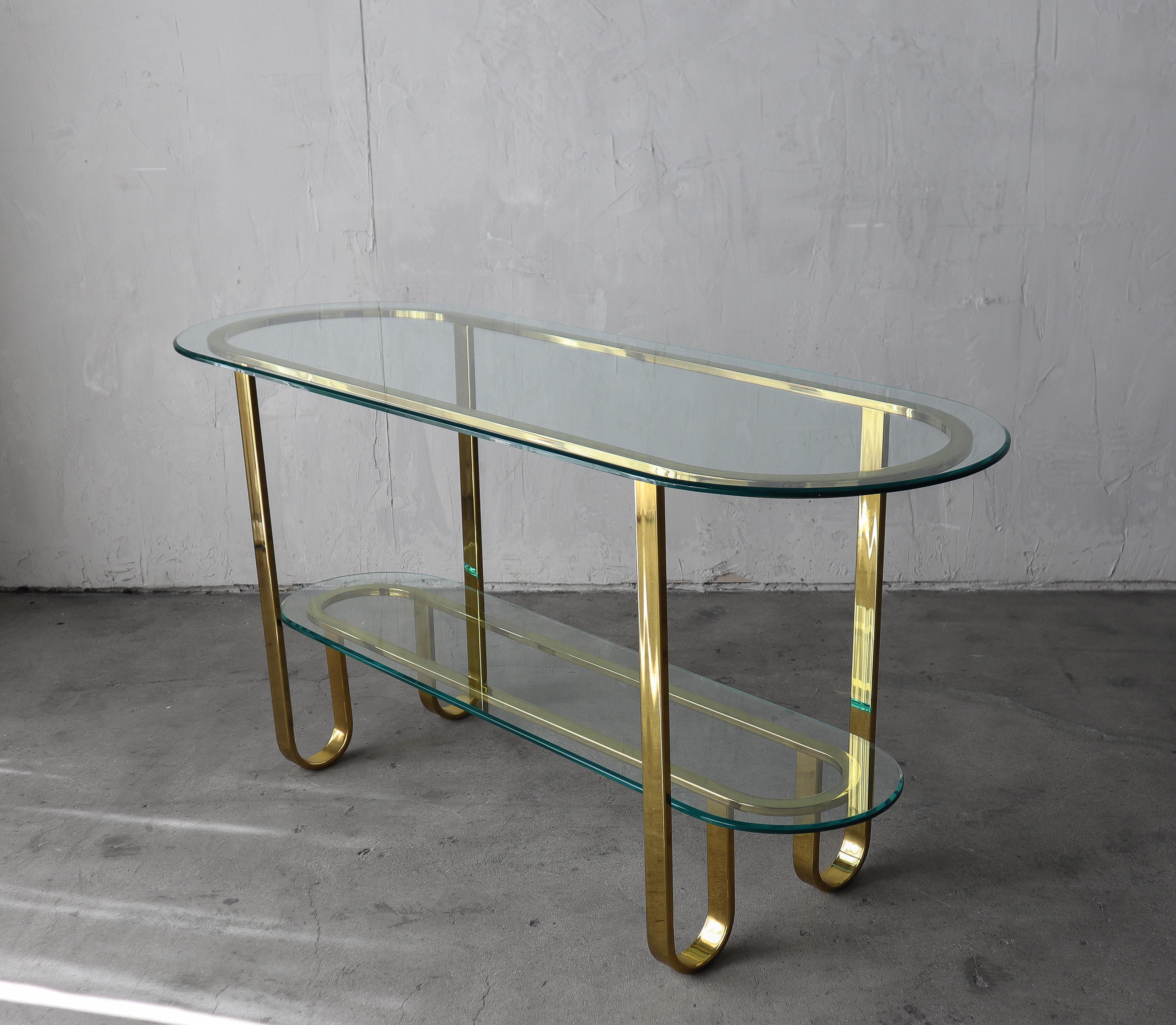 Simplistic yet sophisticated 2-tier brass and glass Mid-Century console table by Milo Baughman. The transparency of this piece makes it perfect in most any space. With two tiers of glass shelving for all your display and storage needs.  It would