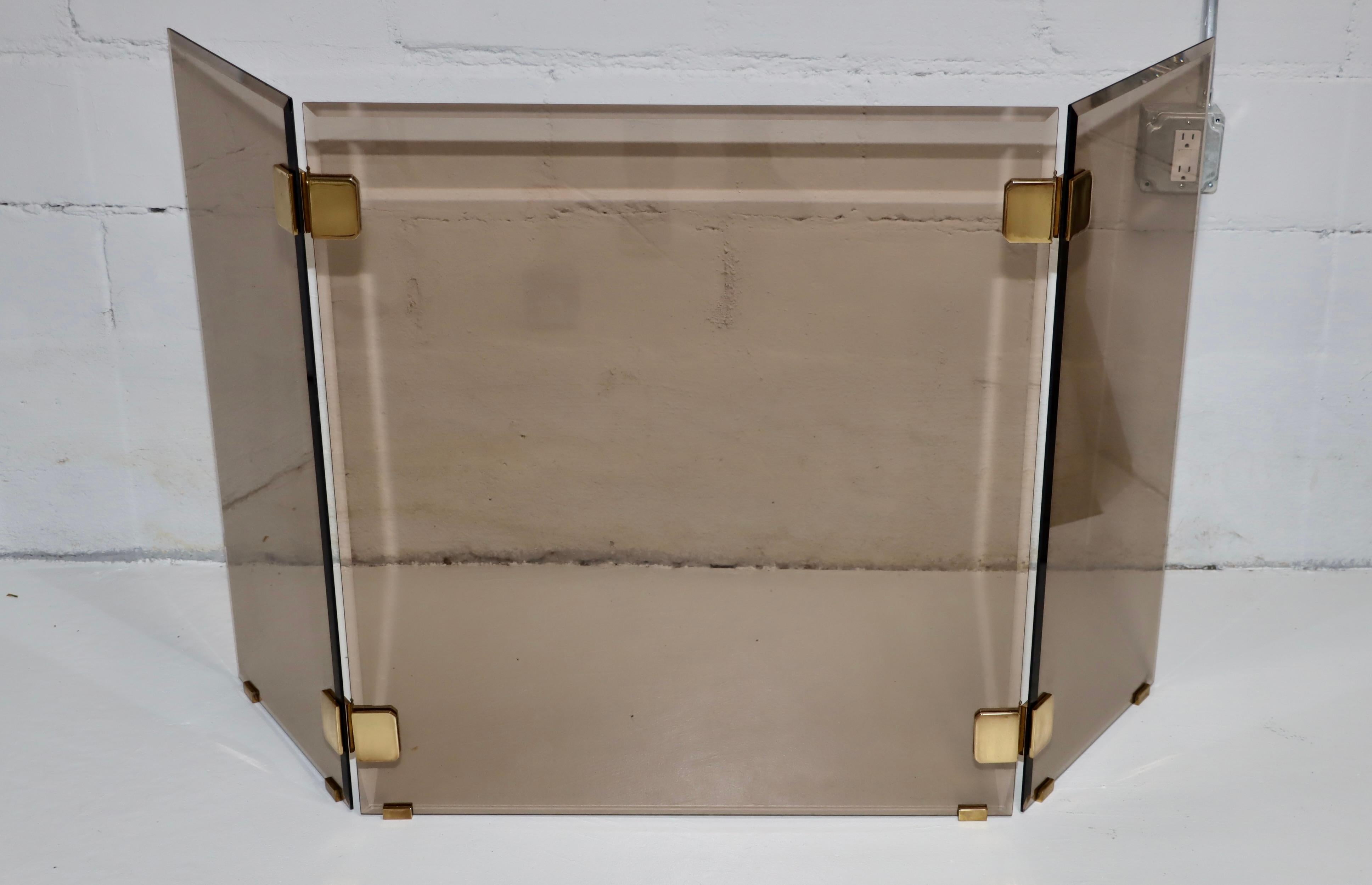 1980's mid-century modern solid brass and smoke glass tri-fold fireplace screen, in vintage original condition with minor wear and patina to the brass due to age and use, there is a minor chip to one of the glass panels as shown in the last picture.