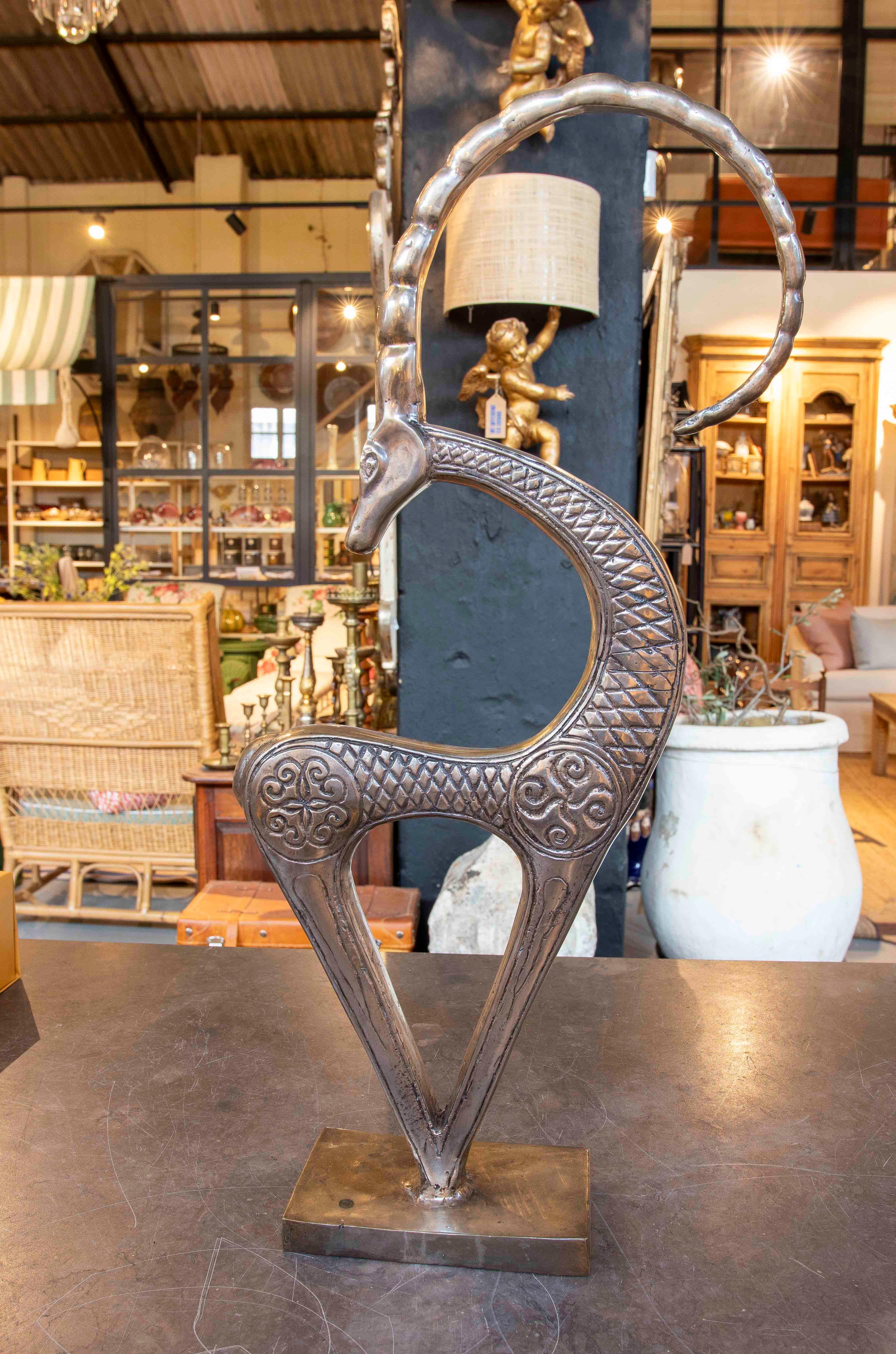 Elegant bronze goat sculpture, with a design that combines stylized lines and ornamental details engraved on its surface. The figure features a contemporary style with traditional influences, highlighting the fluidity of the curves and the detailed