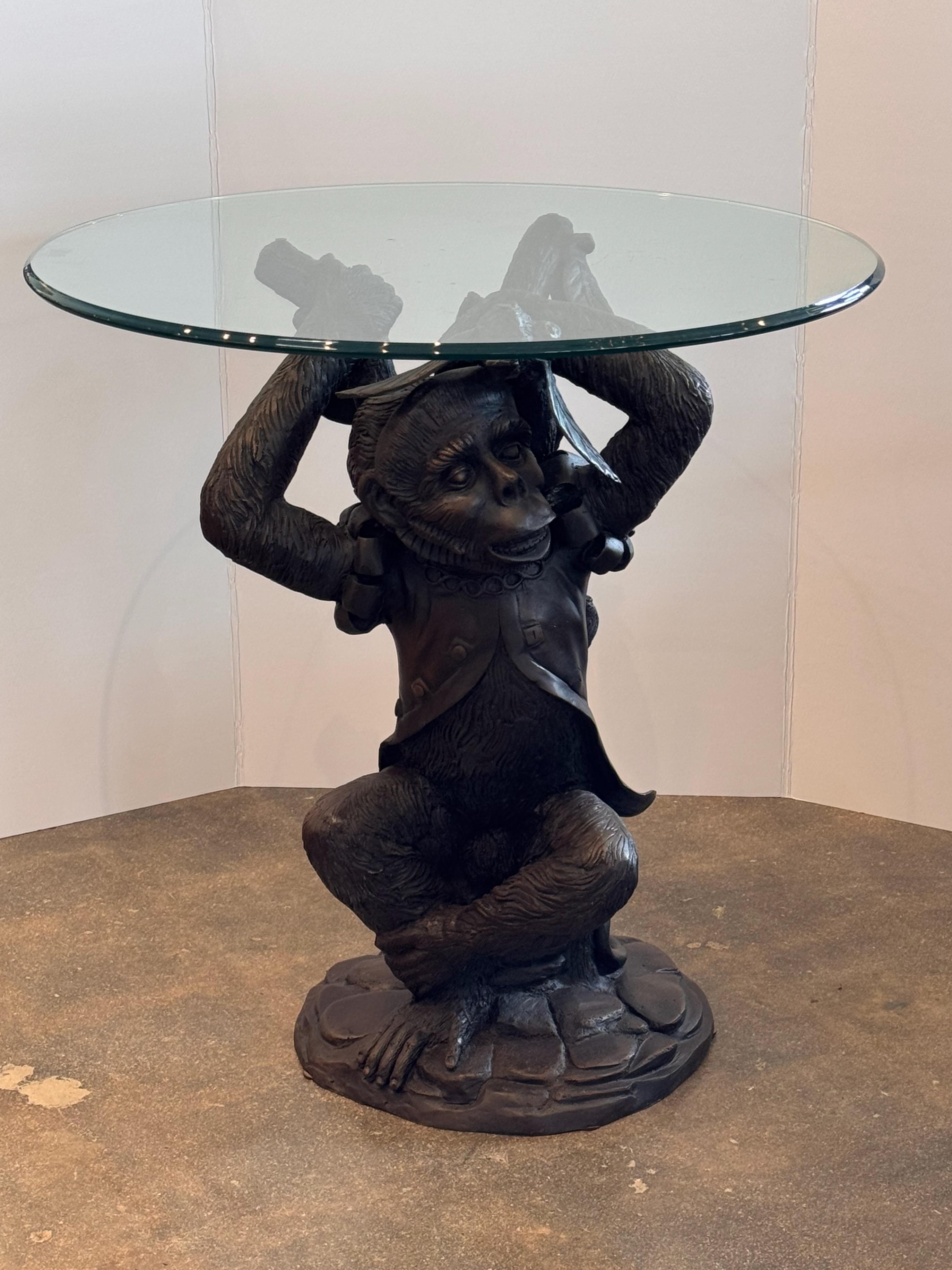 A whimsical bronze monkey table featuring a finely cast figure of a monkey dressed in an ornate jacket, balancing a round glass tabletop on its head. The detailed sculpting showcases lifelike fur texture and expressive facial features, while the