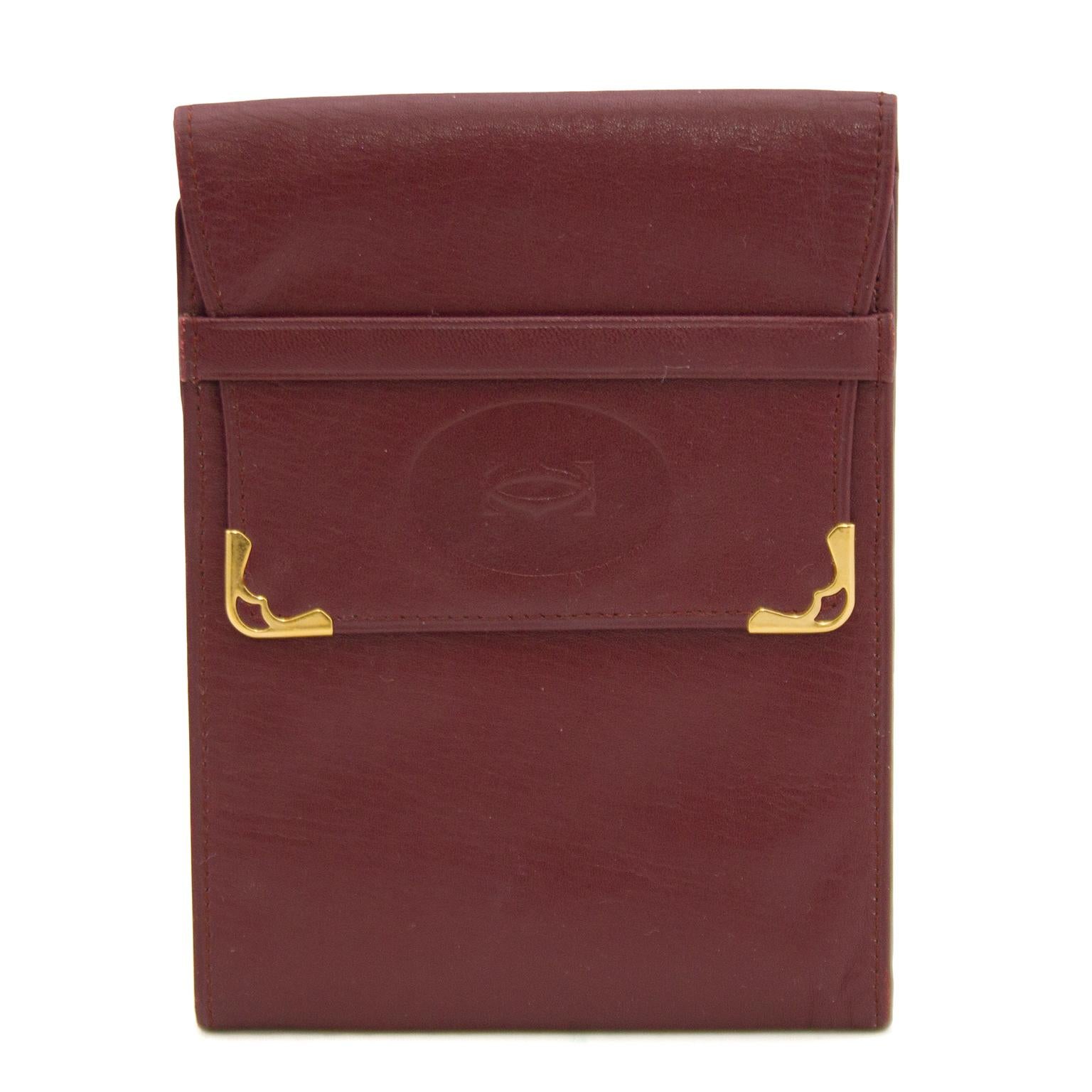Cartier burgundy leather bill fold wallet from the 1980s with a fold over belted closure detail and goldtone metal corners. The flap is embossed with the Cartier CC logo and the entire wallet is stitched in matching burgundy thread. The interior