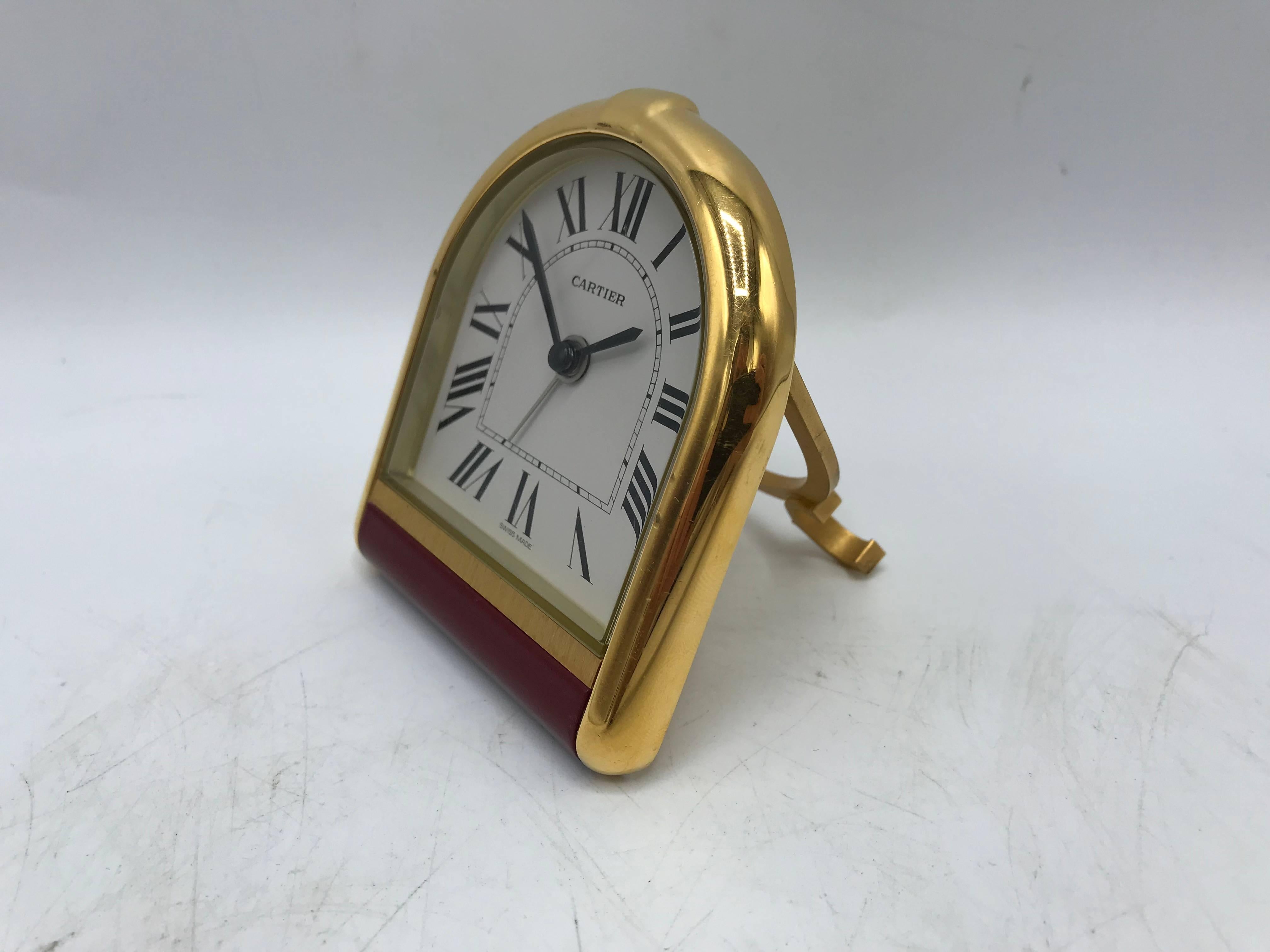 Offered is an exquisite, 1980s Cartier gold travel clock with a red enamel band across the bottom. Glass front. Per Cartier's usual attention to detail, the stand has two mirrored 'C's in Cartier's signature font as the feet, a blue stone on the