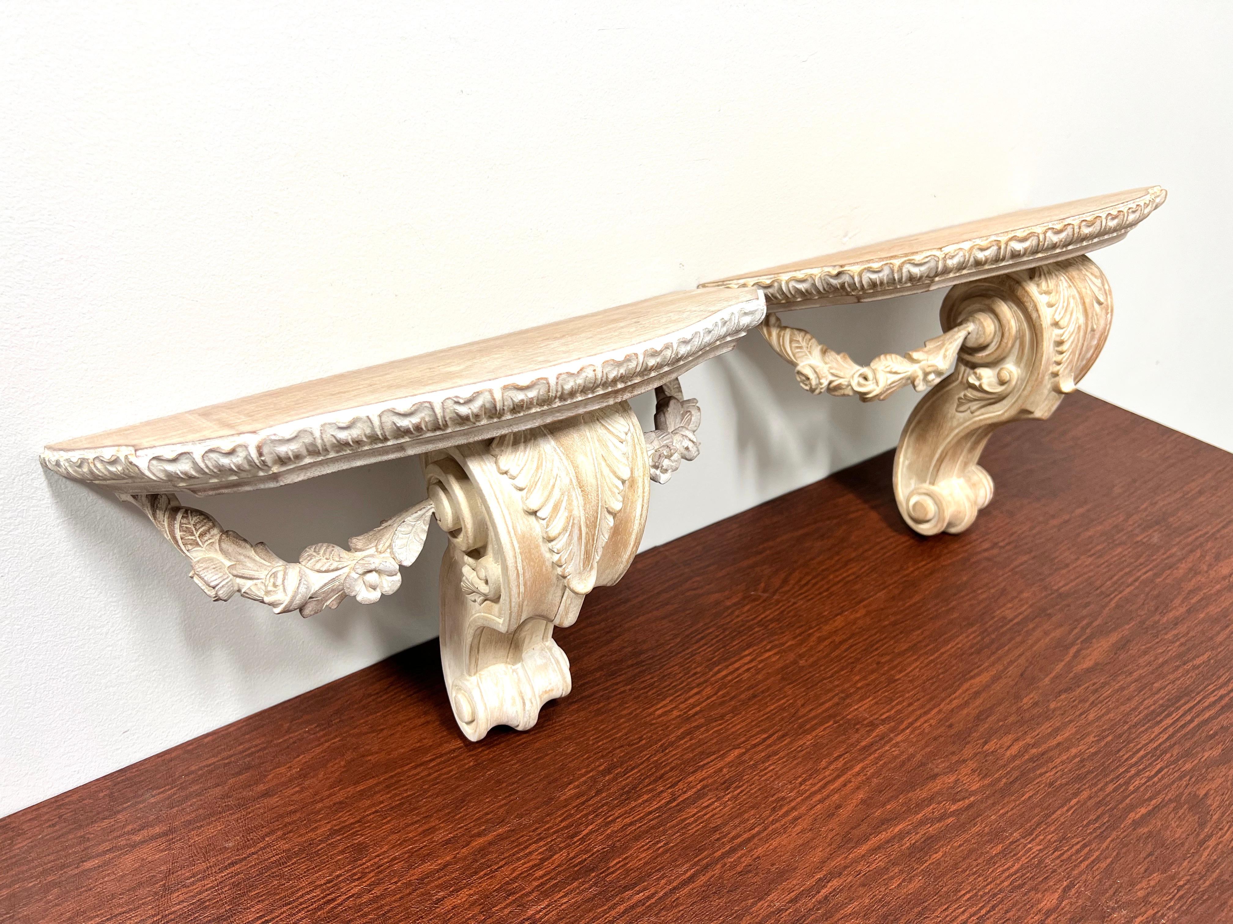 A pair of Rococo style wall bracket shelves, unbranded. Solid wood with a whitewashed finish, flat serpentine shaped display surface, and decorative floral & foliate swags. Dual brass screw holders on back for wall hanging. Made in the USA,circa