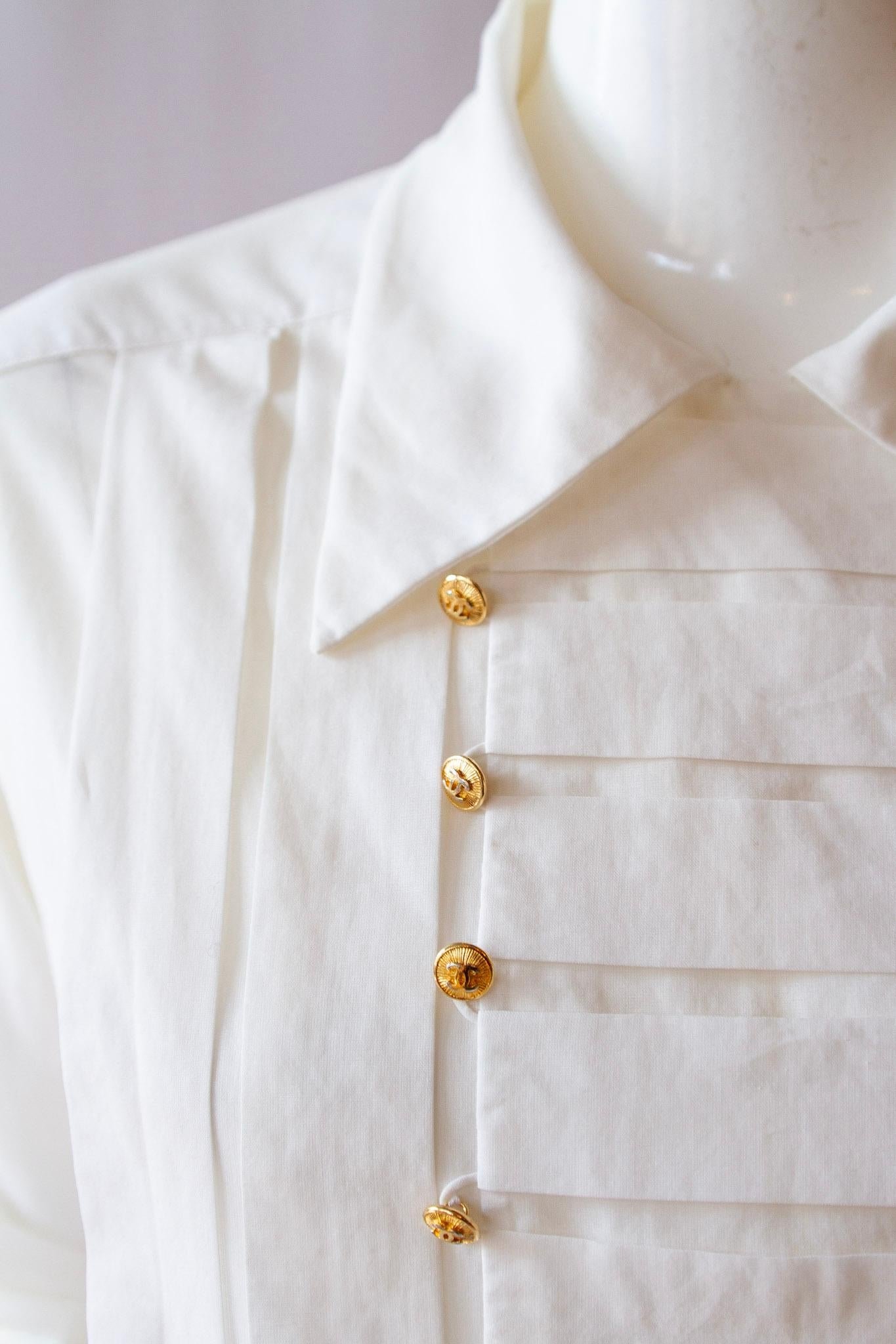 Top à col à manches longues en coton blanc de Chanel, style smoking, avec panneau plissé sur le devant, orné de boutons Chanel dorés.