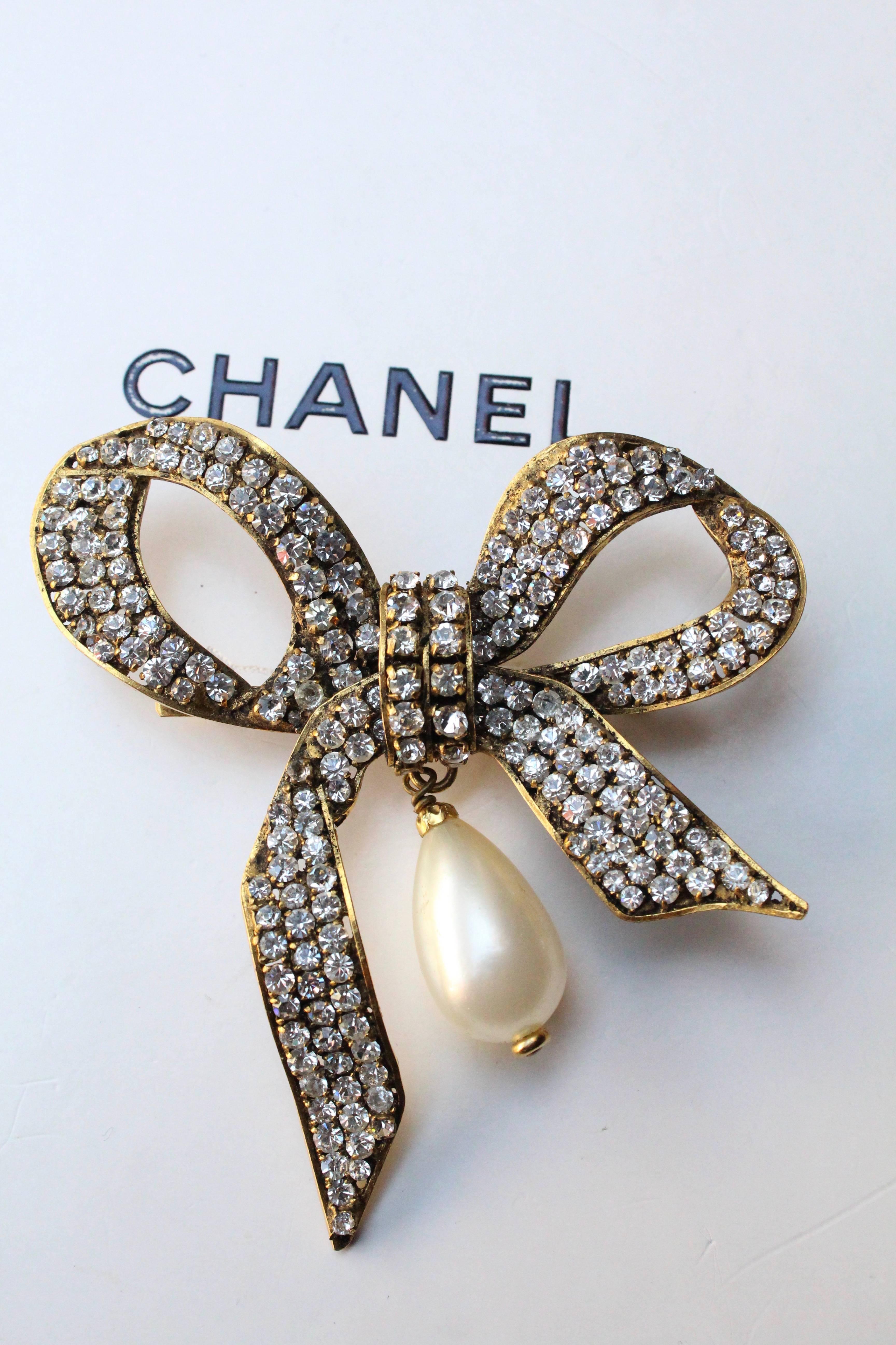 CHANEL (Made in France) Lovely large evening brooch in the shape of a bow, composed of a gilded metal setting entirely paved with white rhinestones. The brooch is further embellished with a pearly glass tear drop.

Signed on a back plate.

Circa