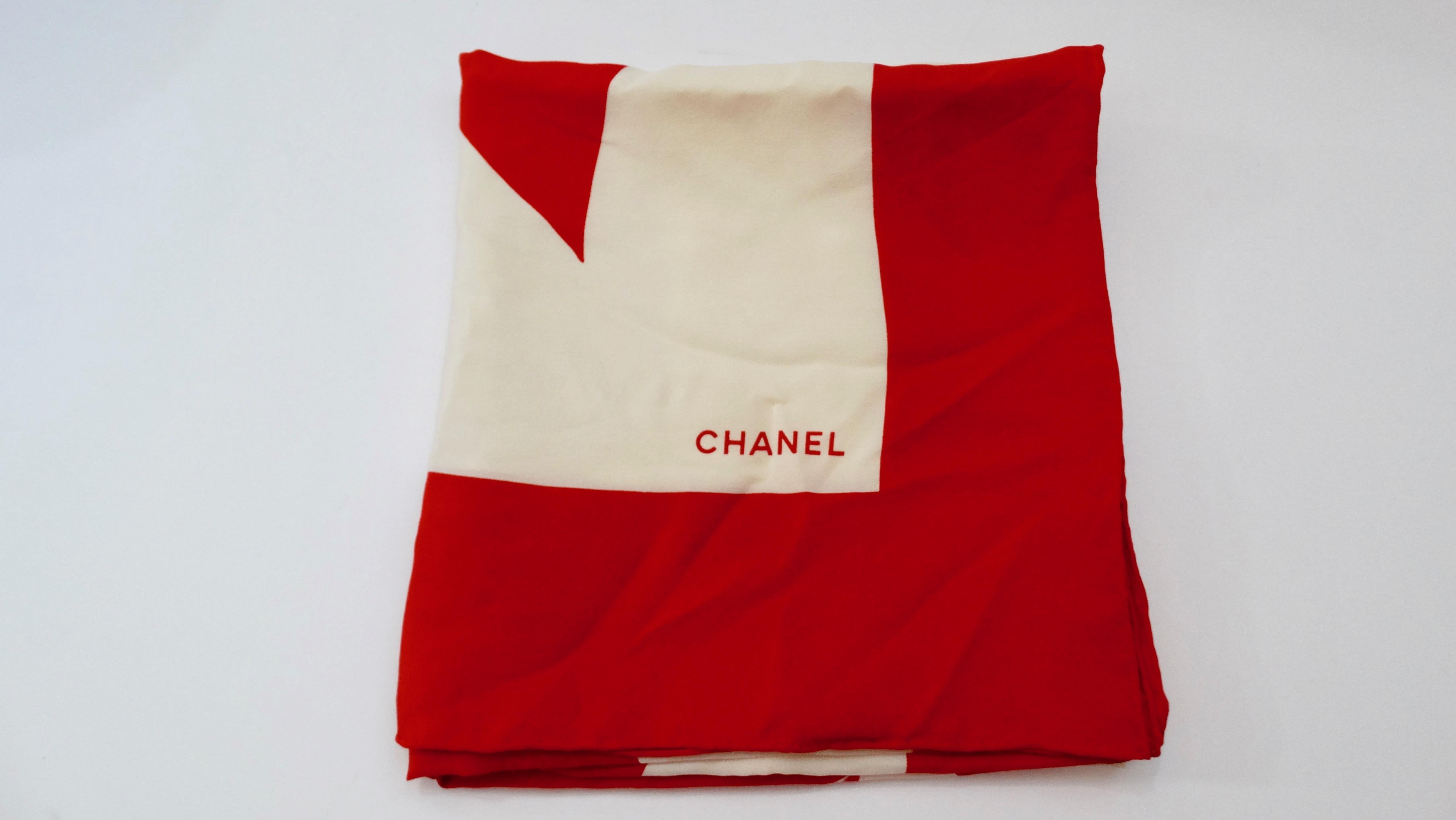 This scarf features the signature Chanel Camellia flower in creme-white and lipstick red in the center of a red bow on a creme-white background with a red border. It is made from crêpe de Chine, has hand-rolled edges and is signed 