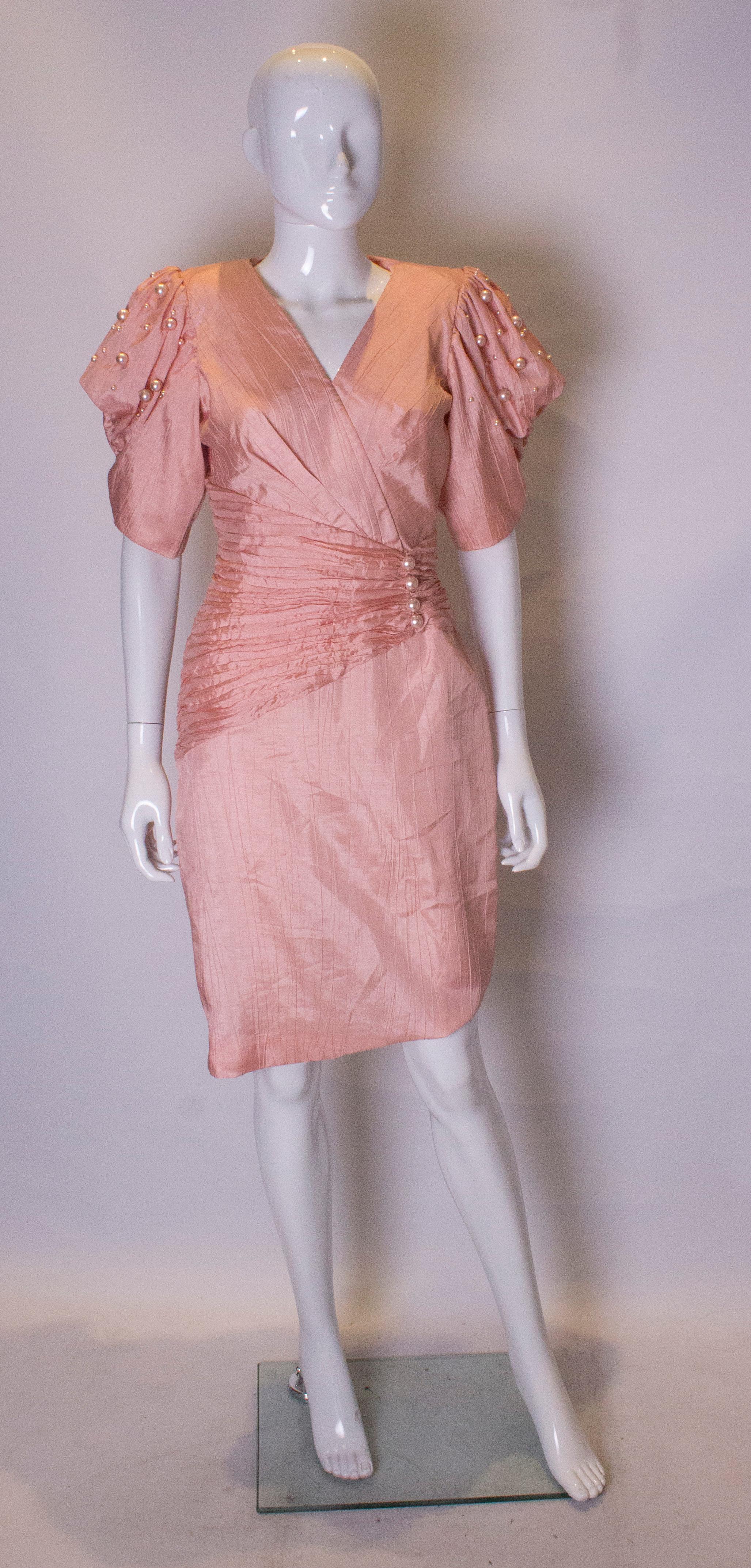 A head turning cocktail dress in a pretty powder pink colour with pearl decoration on the sleeves and waistband. It has a wrap over v neckline and asymetric waist band. It has a central back zip and gathering on the shoulder.
