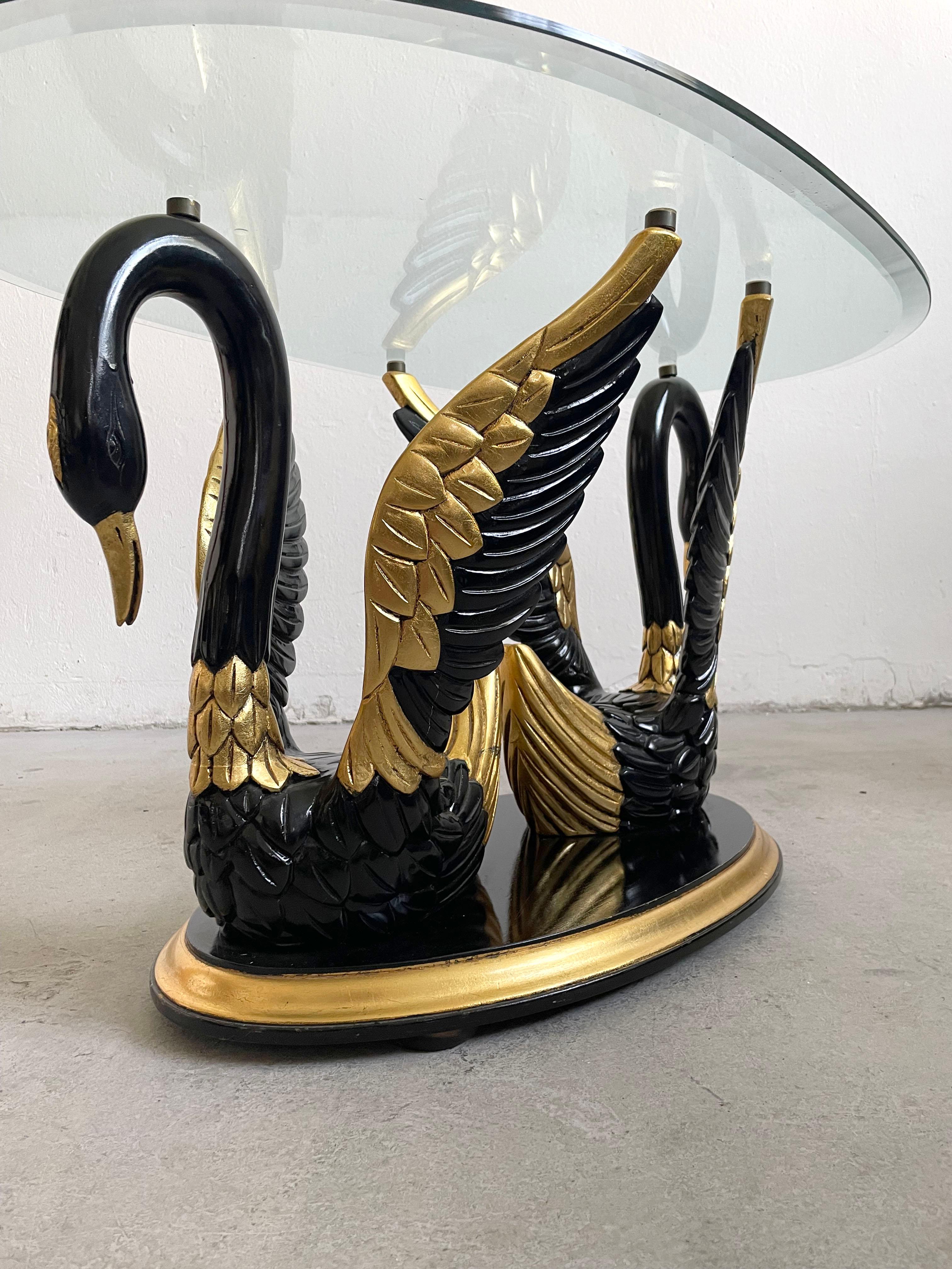 A Hollywood Regency style from the 1980's, a guilty pleasure for design lovers, the coffee table featuring a pair of swans carved in wood, painted in glossy black and gold.
This sculptural table has a free standing clear faceted oval glass top.

The