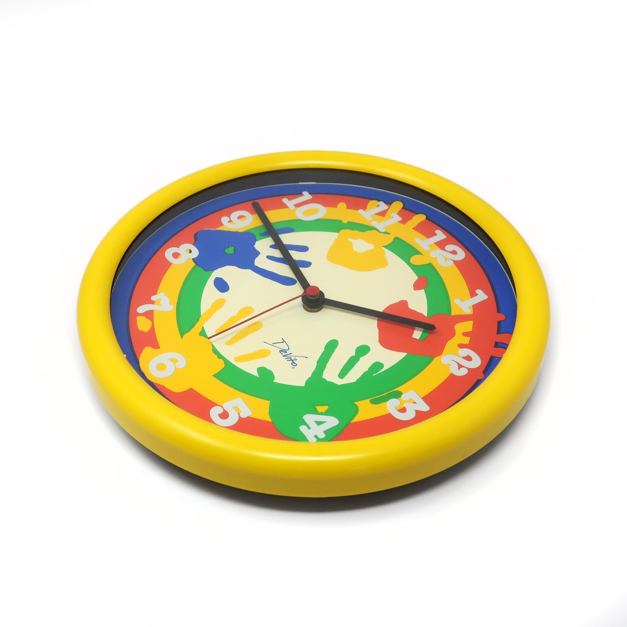 A fun postmodern colored wall clock from the 1980s by Devito.  It has a yellow plastic case, blue, red, yellow, white and green face with multicolored handprints, and black and red clock hands.  Brings a lot of color to any room, whether it be a