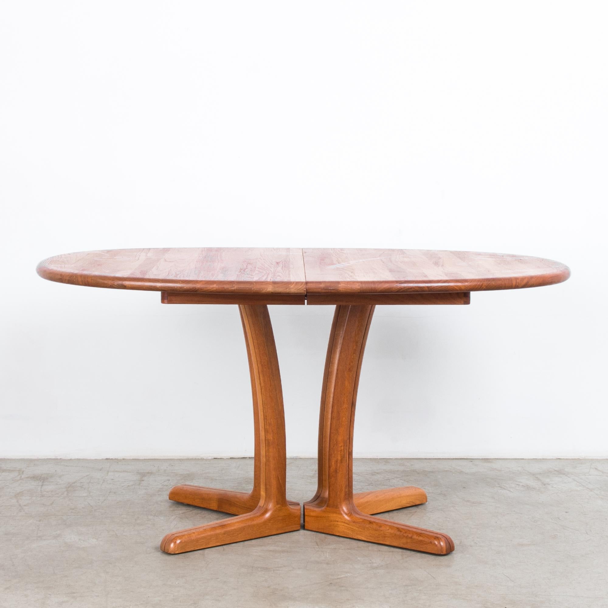 A teak dining table from Denmark, circa 1980. A round tabletop sits atop four legs with distinctive splayed feet. An extra leaf allows the tabletop to be extended to create a long, oval dining table. The wood is a bright caramel color, with the
