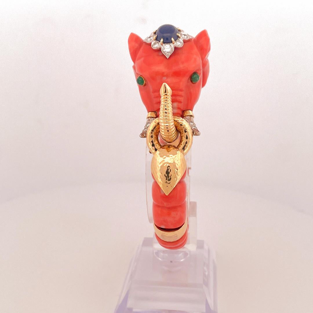 From the designer David Webb circa 1980’s, 18 karat yellow gold coral and gemstone elephant bangle. This bangle is crafted with carved coral in the shape of the elephant with an oval cabochon cut blue sapphire weighing approximately 2.68 carats on