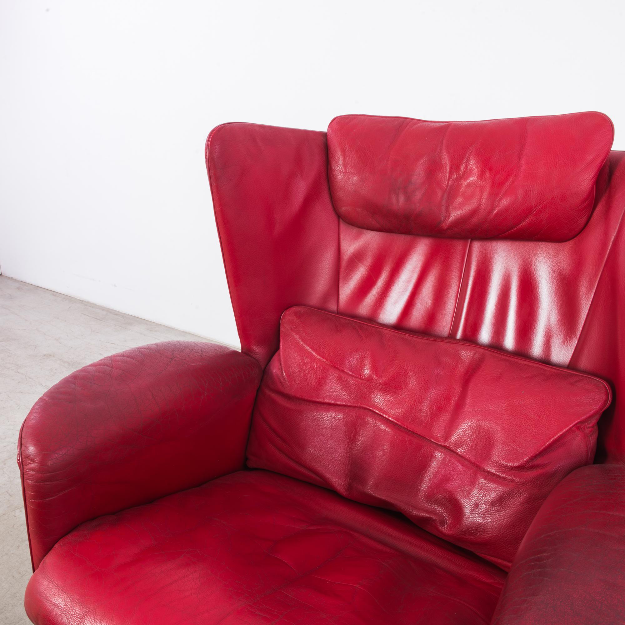 A leather armchair by Swiss furniture designers De Sede, circa 1980, with accompanying ottoman. The leather is a vivid crimson, with the silky finish of an expensive lipstick. The high wings and full curve of the backrest create a dramatic