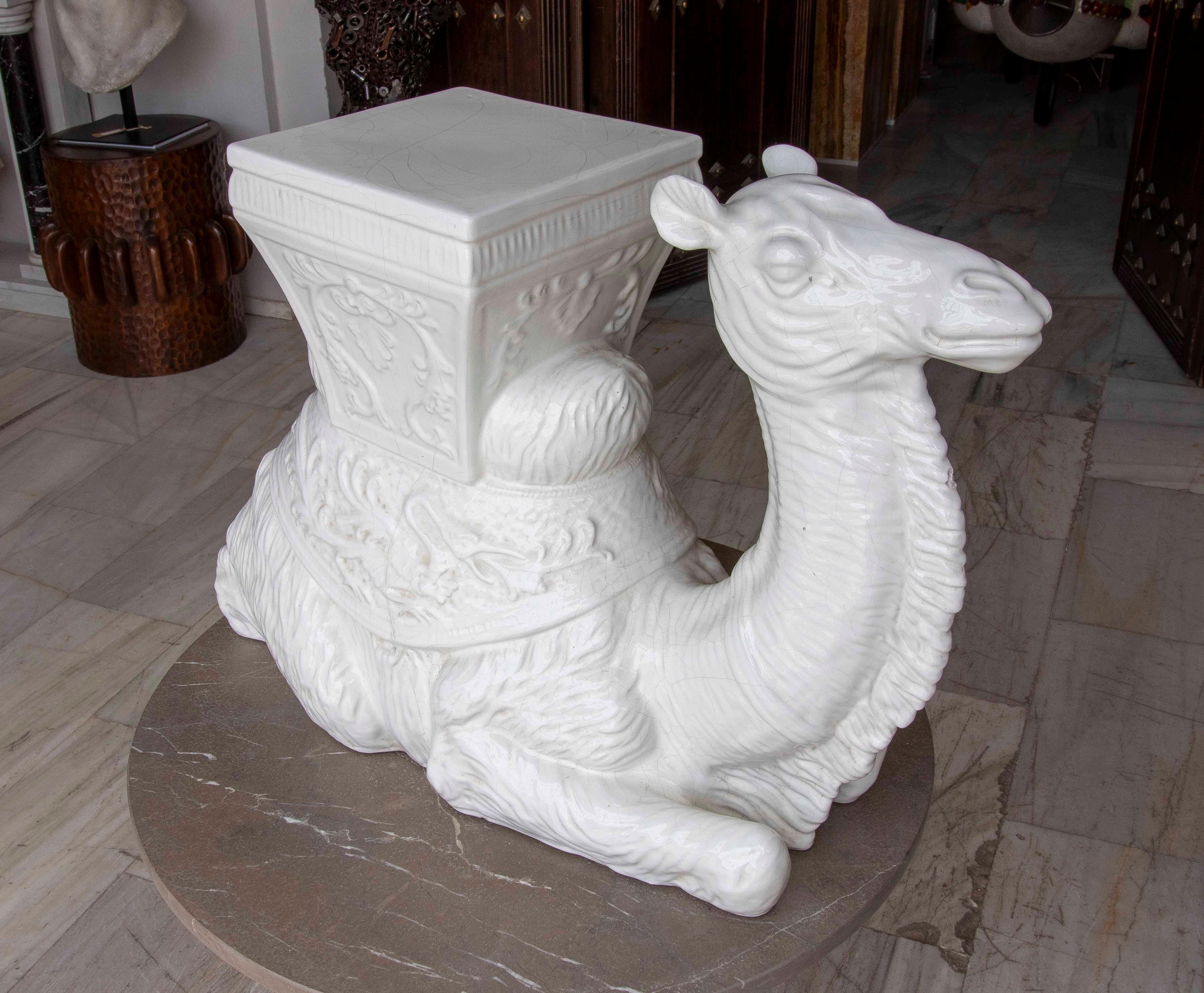 An original reclining camel sculpture made of pure white glazed ceramic, with a glossy and elegant finish. The figure represents a reclining camel, intricately sculpted, with an ornate mount serving as a flat base, ideal for use as a side table or