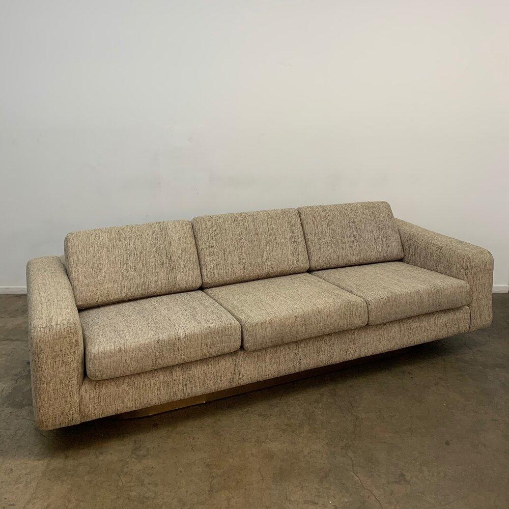 Measures: W 95 D 36.5 H 22.5 

Vintage sofa in the manner of Milo Baughman. Item is in great gently used condition with no large areas of wear, minor dents in brass pictured. Item offers deep seating and sits on a wood plinth wrapped in brushed