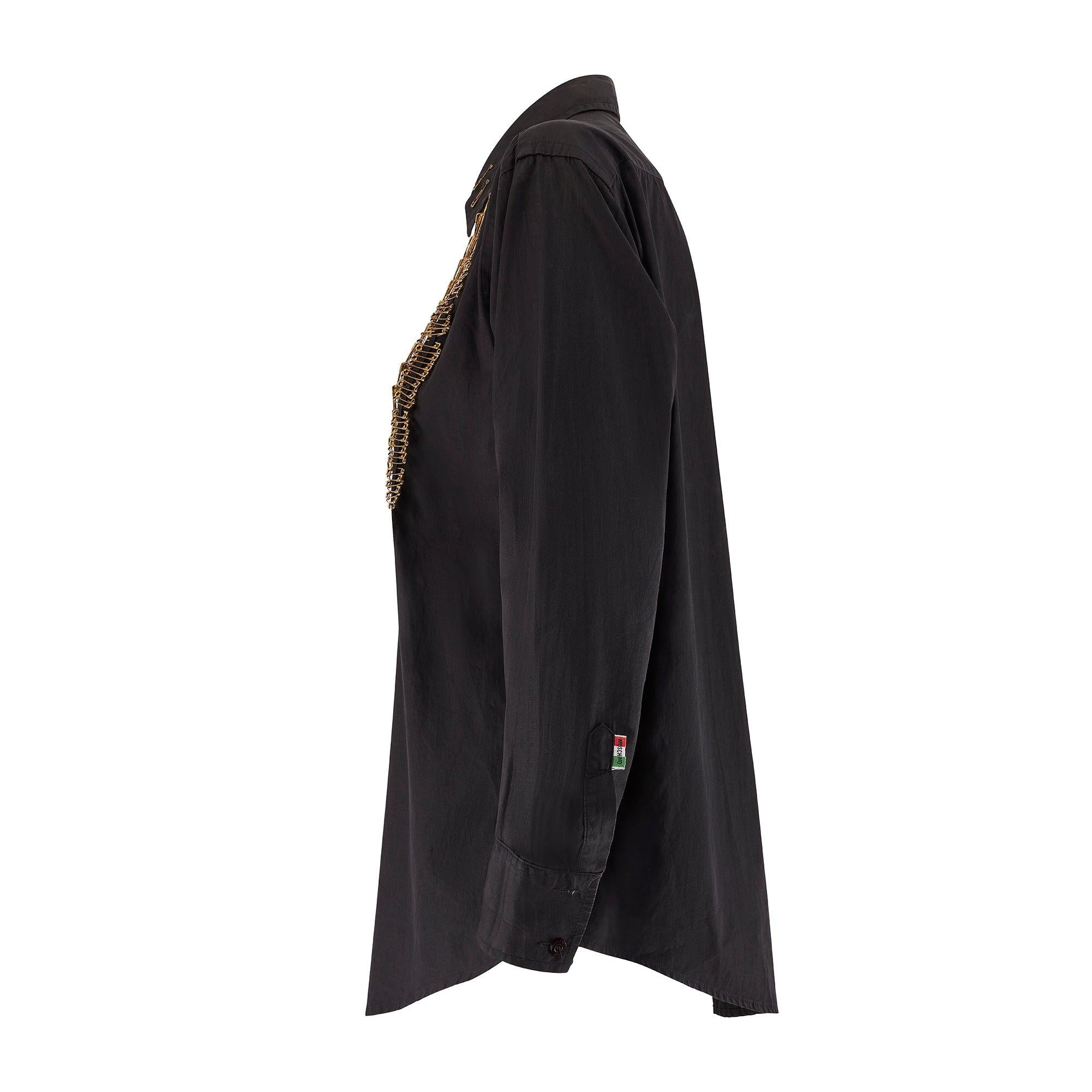Realizzata in cotone nero, presenta una silhouette allungata con spacchetti sul retro per facilitare i movimenti. I polsini sono lunghi e sono fissati con due bottoni marrone scuro con inciso il nome del marchio. La parte anteriore della camicia è