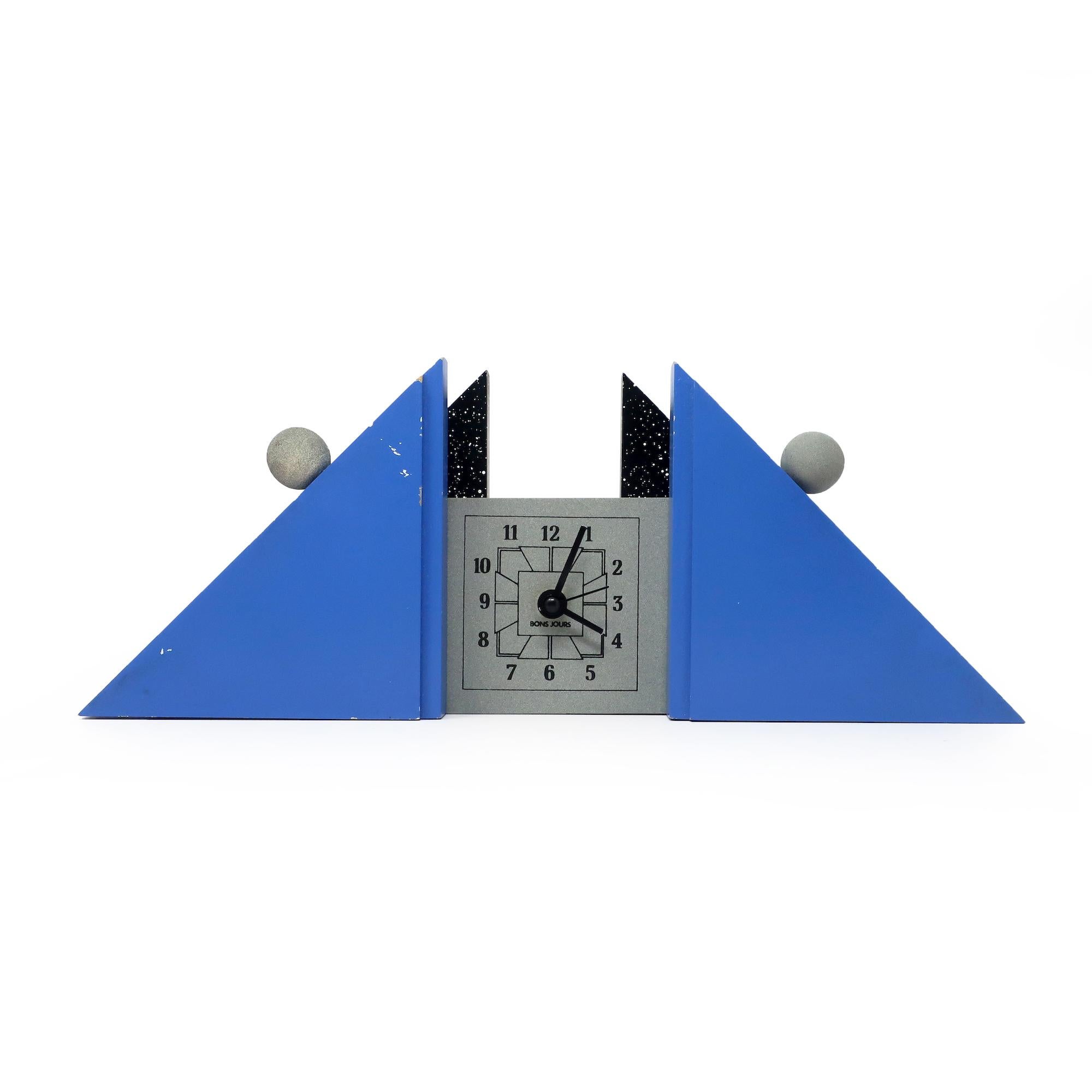 A sculptural French postmodern mantel clock with blue and splatter painted supports, gray face and accents, and black numbers and hands.  A fantastically contrasting low triangular shape with round details and a beautifully three dimensionality. 