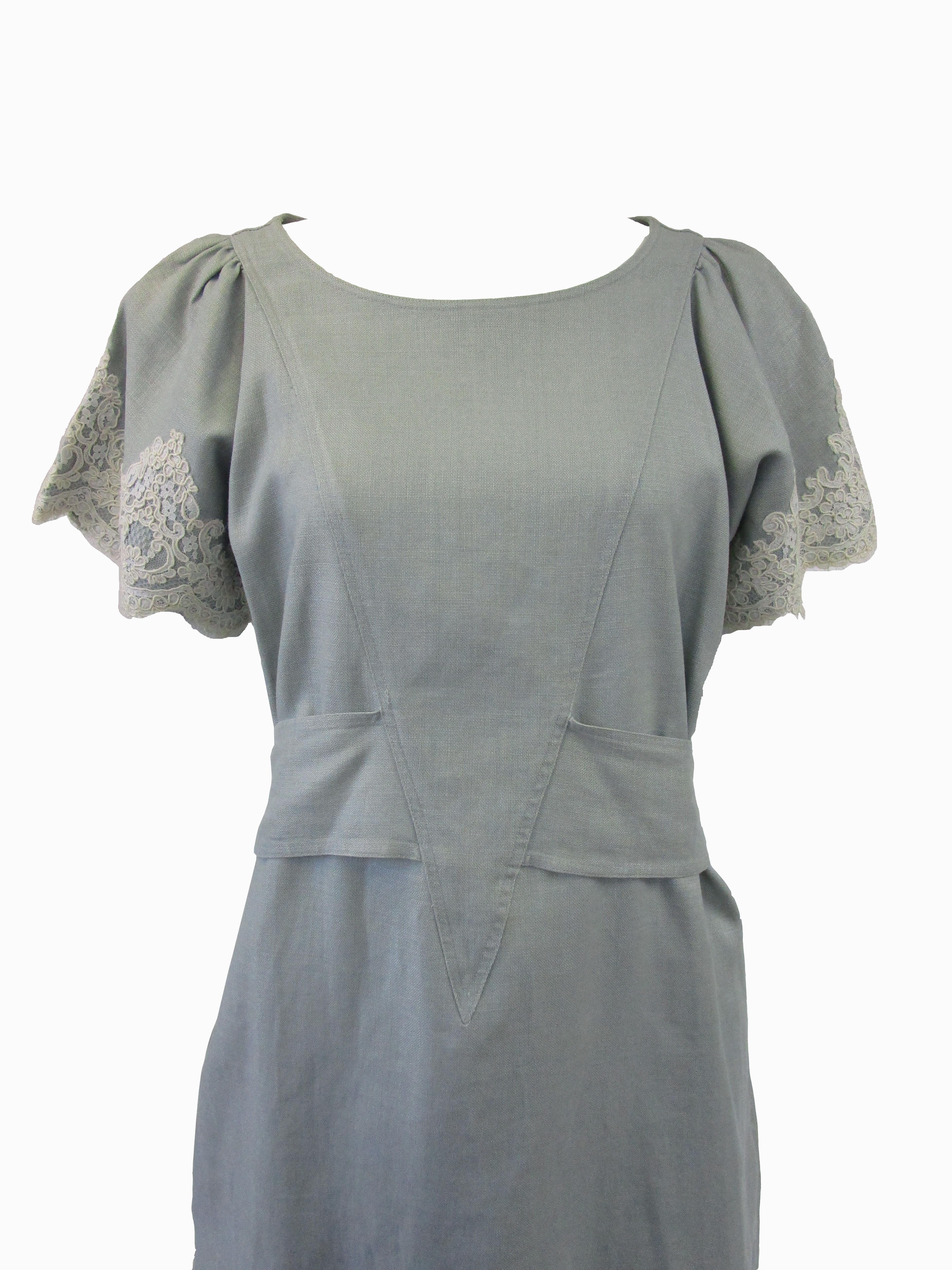 
Lovely day dress from Geoffrey Beene! This dress features a dusty miller colored linen fabric in a shift silhouette with a structured tie around the waist that can be adjusted to the wearer. It is accented by beautiful bridal lace details around