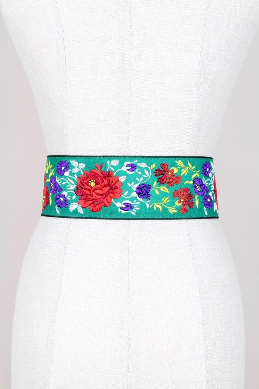 This is a pretty wide waist belt by French designer Georges Rech from the 1980s. Made of viscose fabric and suede leather, it features an embroidered floral motif in red, purple, white and yellow on a bright green background and closes with a