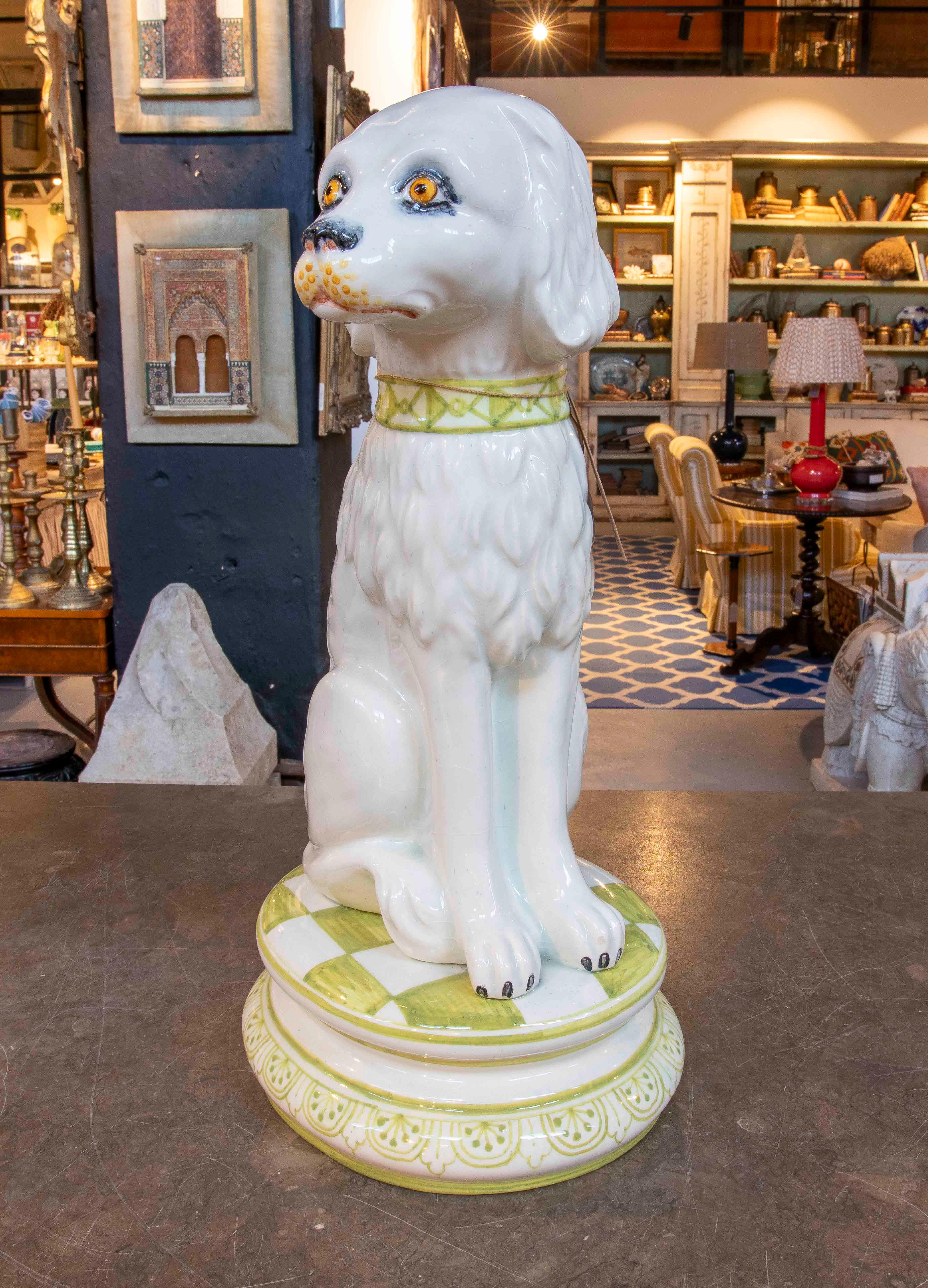 Charmante sculpture de chien en céramique émaillée blanche, avec une expression sereine et des détails peints à la main sur le museau et les yeux qui lui donnent une touche de caractère. La pièce repose sur une base circulaire avec un motif