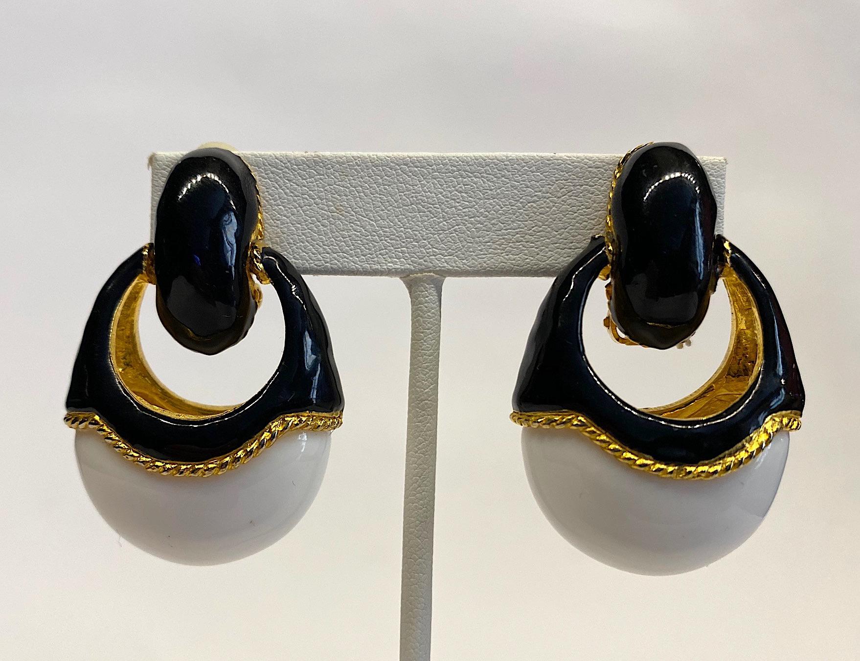 A stylish and chic pair of late 1980s door knocker style dangle earrings. Each clip earring is gold plated with black enamel on top and a large oval white lucite cabochon set into the bottom. The interior of the earrings and rope border around the