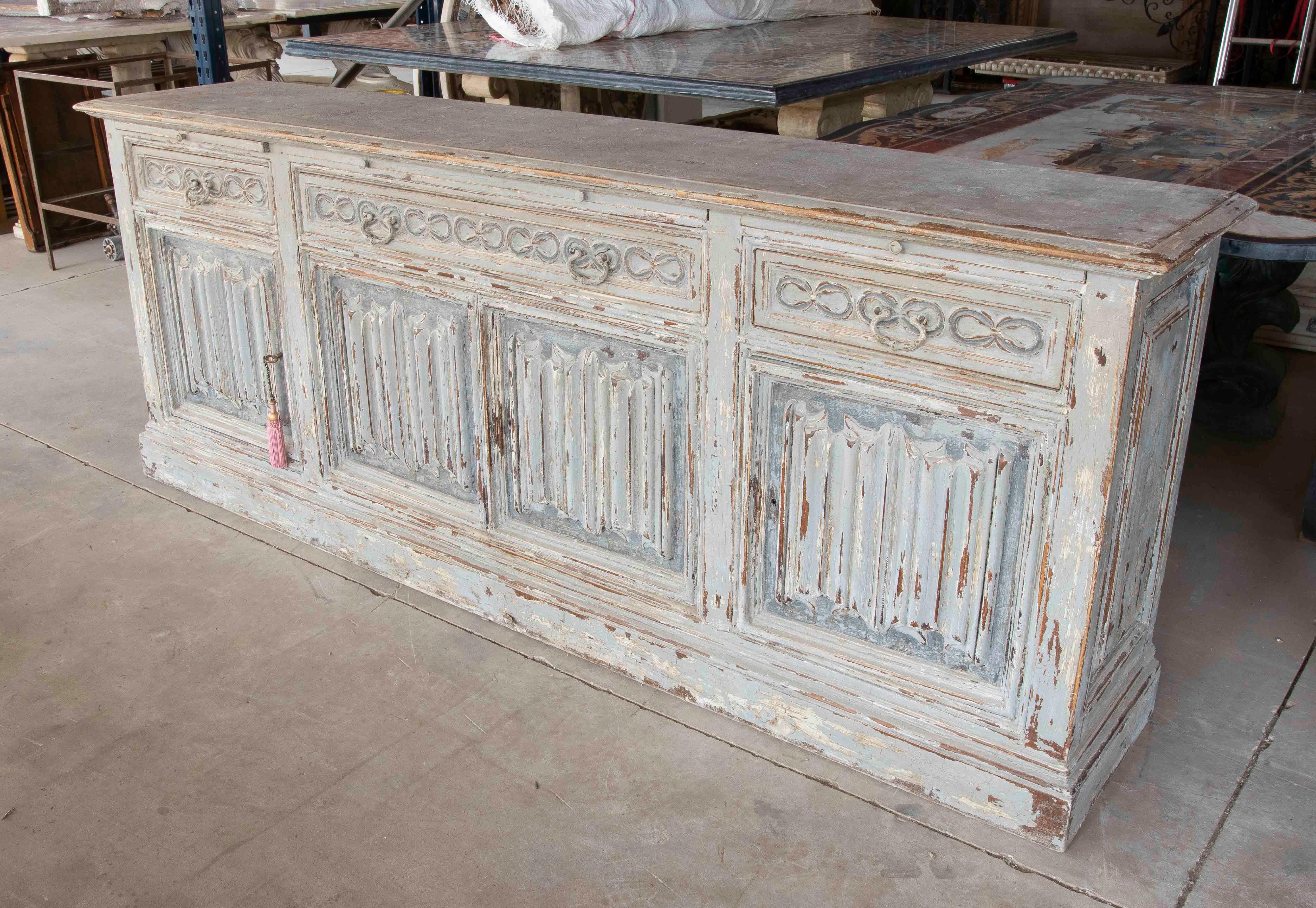 Elegant Provençal-style sideboard, crafted from solid wood with a polychrome finish in white and gray tones that enhance its antique patina. It features four carved doors with fluted vertical reliefs and ornamental floral details on the upper