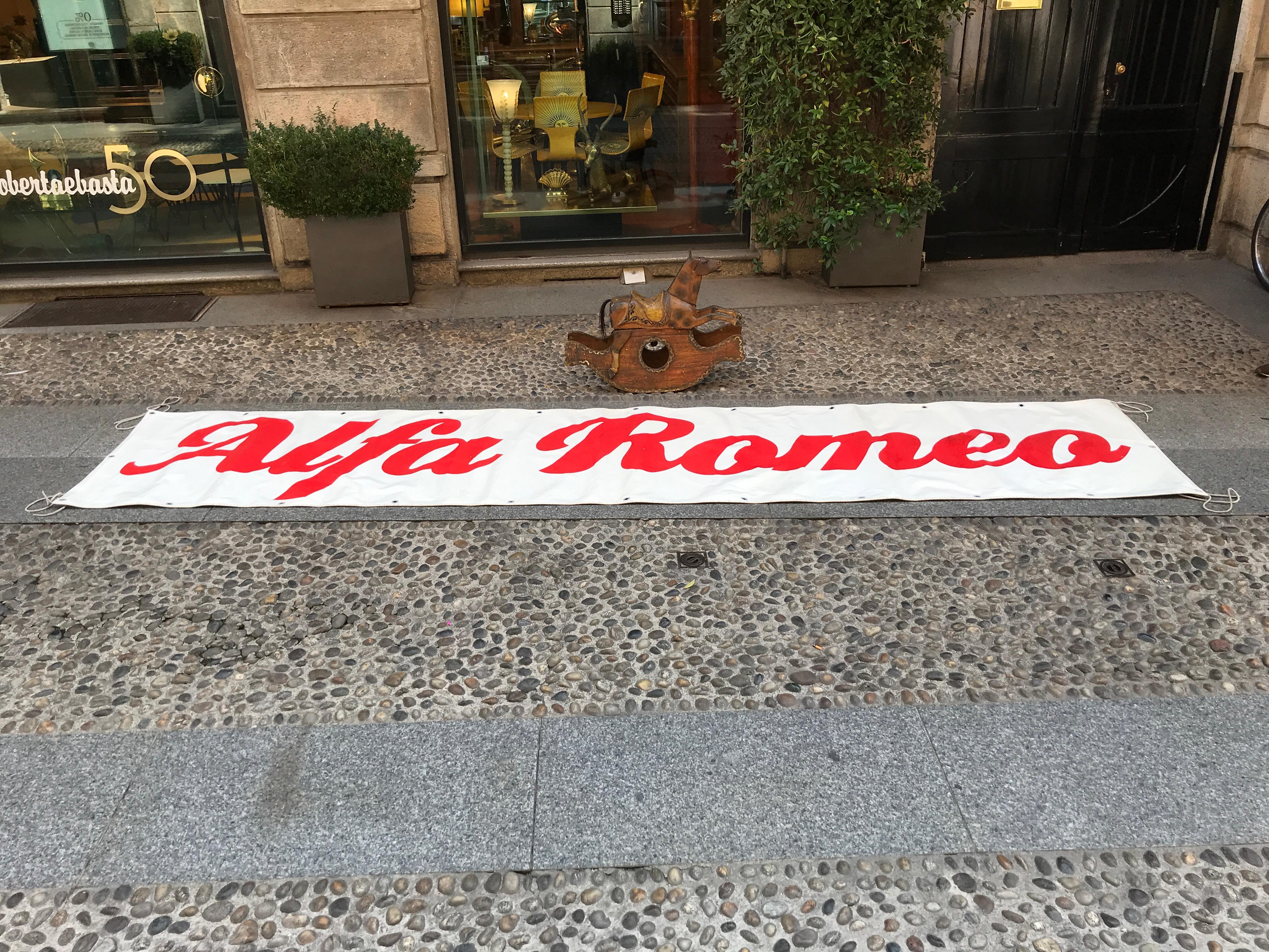 Vintage large rectangular Alfa Romeo PVC banner produced in Italy in the 1980s.

The red Alfa Romeo logo is on a white background. The banner is framed by multiple eyelets, and also four nylon ropes at each corner.

Collector's note:

Alfa