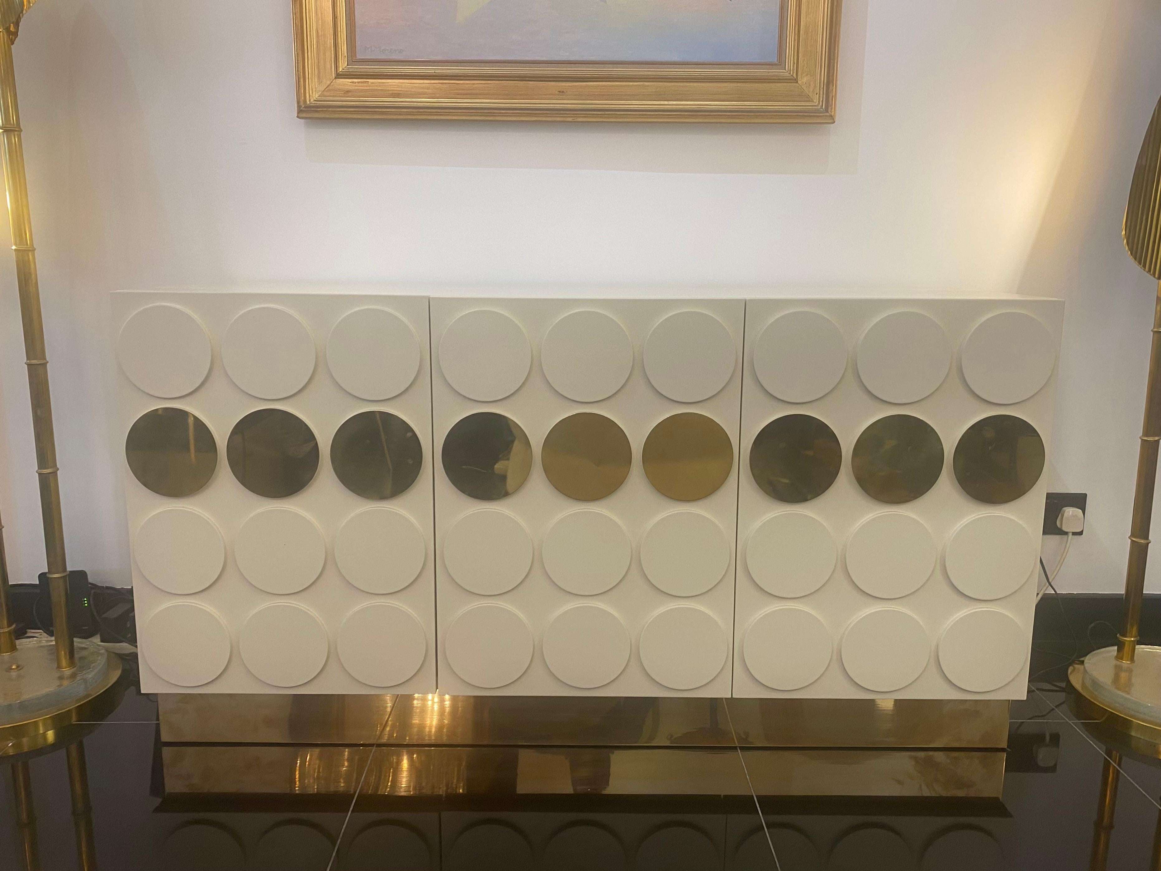 An Italian cabinet 3 door cabinet designed with raised circles in white pickle lacquer and brass on all three sides. There is a single shelf in natural wood on both sides with brass piping. The cabinet stands on a glamorous brass surround base.