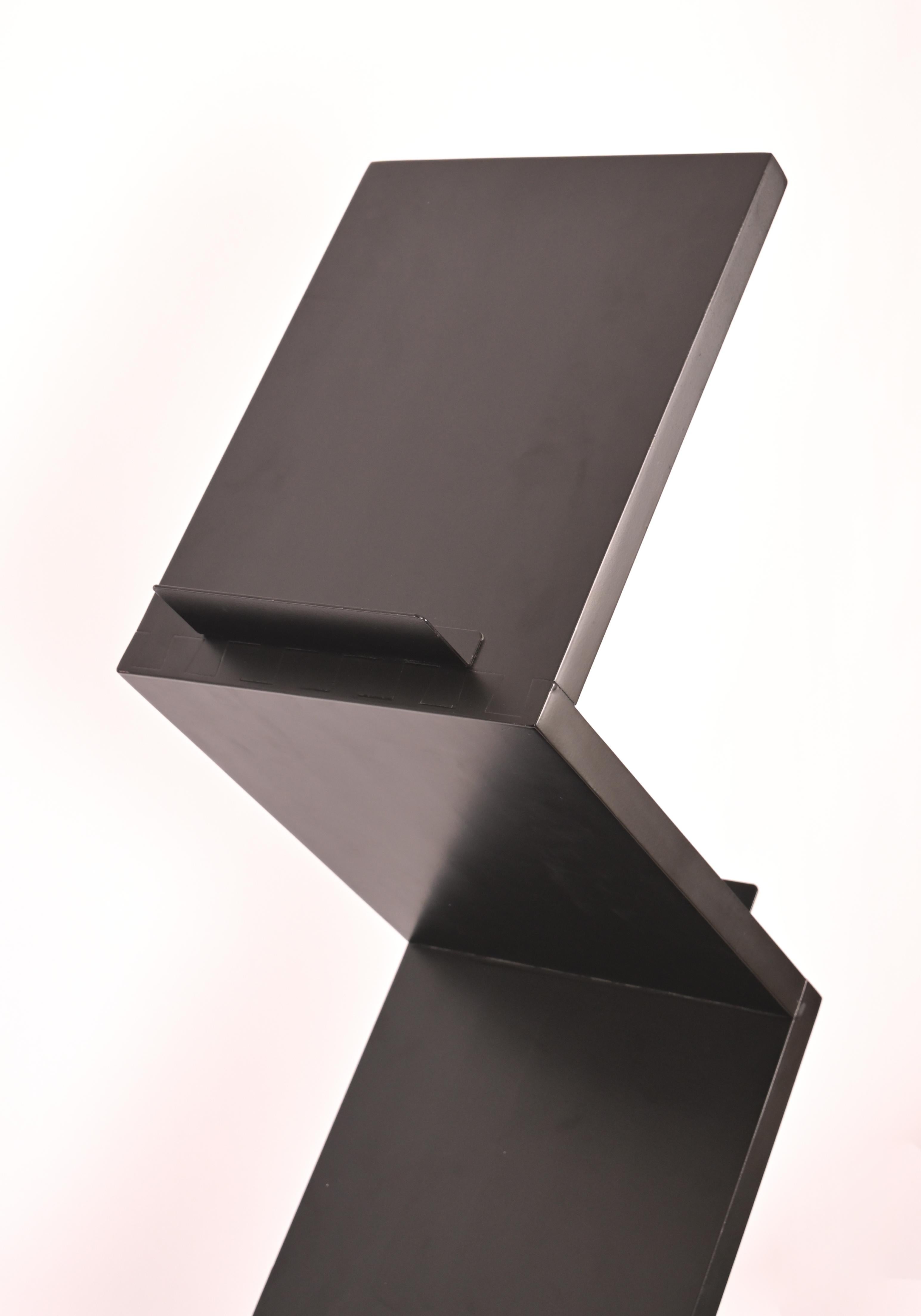 A zig zag shaped vertical display vehicle designed for books with three black painted shelves at a 90 degree angle. The black painted wooden frame is supported by cast concrete base. Designed by Swedish creator Jonas Bohlin ( 1953-). Holds 6 spaces