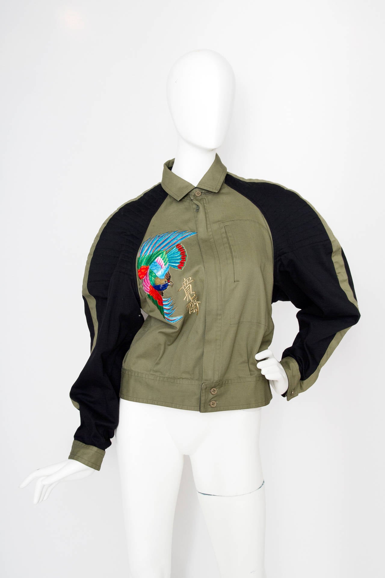 A 1980s Kansai Yamamoto cropped cotton bomber jacket with an extravagant embroidered bird pattern placed both on the front and on the back. The army green jacket has black panels on the sleeves and linear quit detailing on the shoulders. The jacket