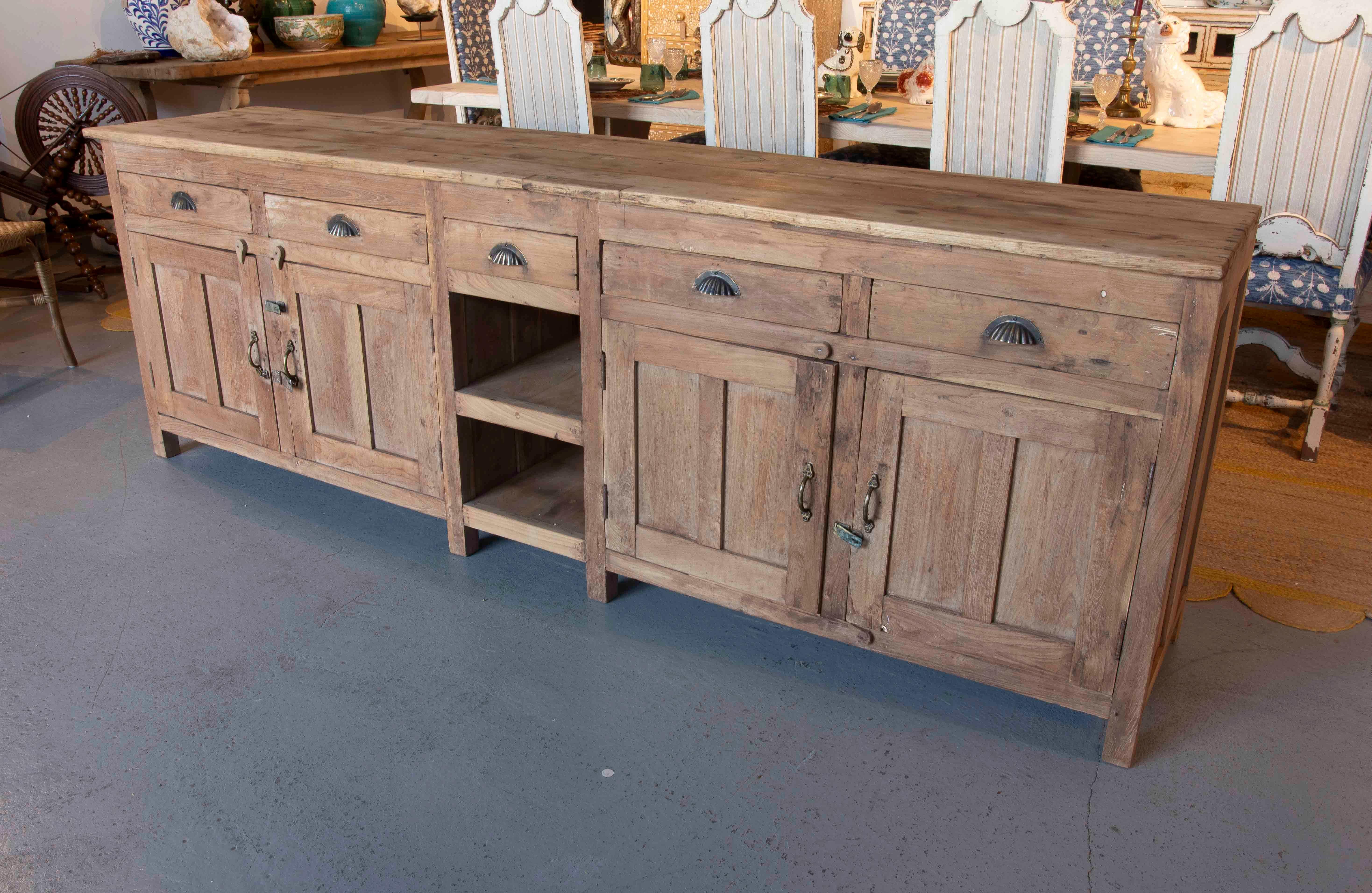Ein beeindruckendes rustikales Sideboard aus Massivholz, das durch seine großzügigen Abmessungen und seine schlichten Linien die Eleganz des Materials unterstreicht. Das Möbelstück verfügt über vier obere Schubladen mit Metallgriffen im Muschelstil,