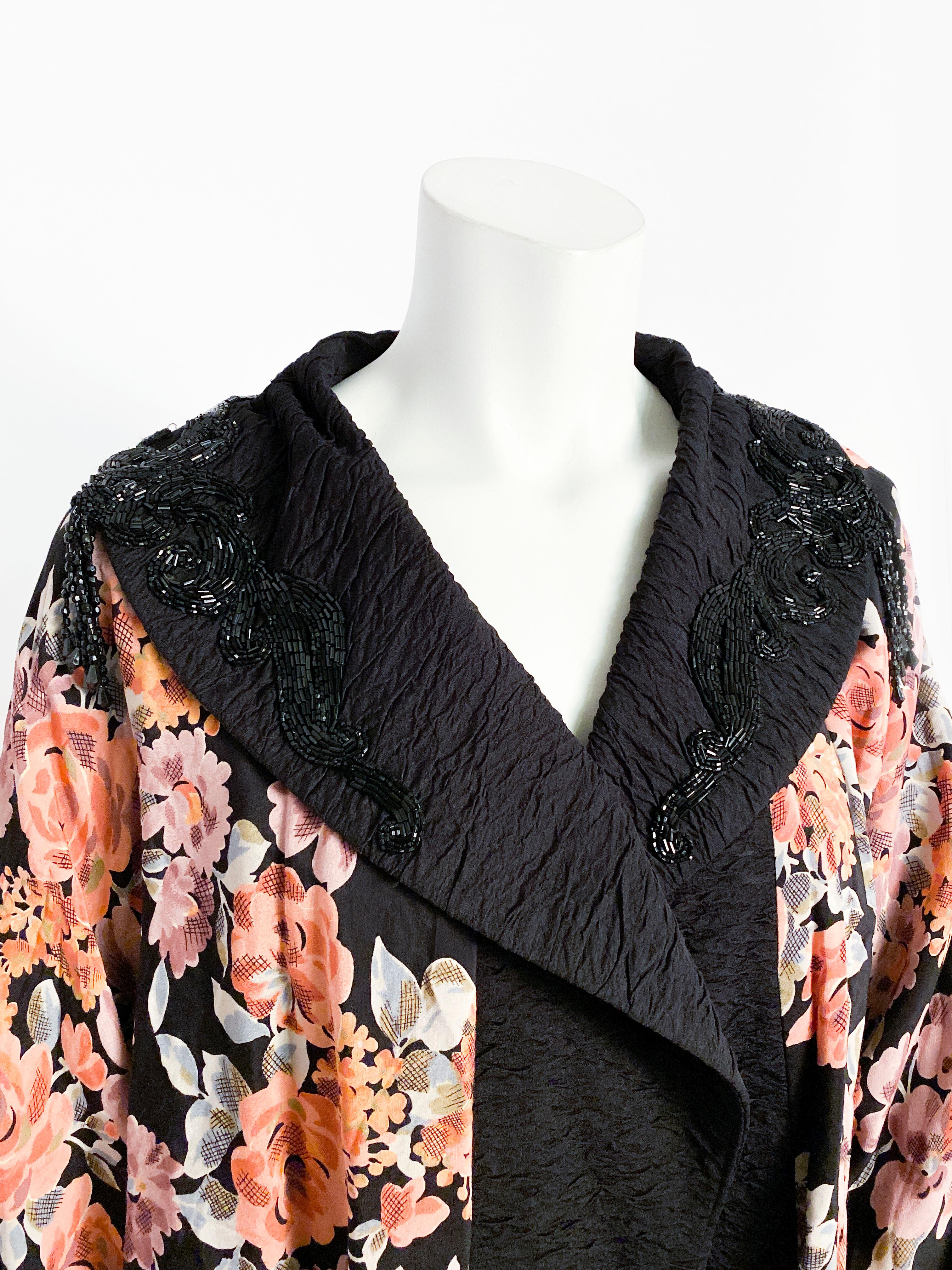1980s Lorrie Kabala cocoon coat featuring a vibrant floral print to contrast the black shawl collar and border all made of a crinkled nylon. The collar is adorned with large jet black beaded applique and a panel of fringe on the back of the neck.