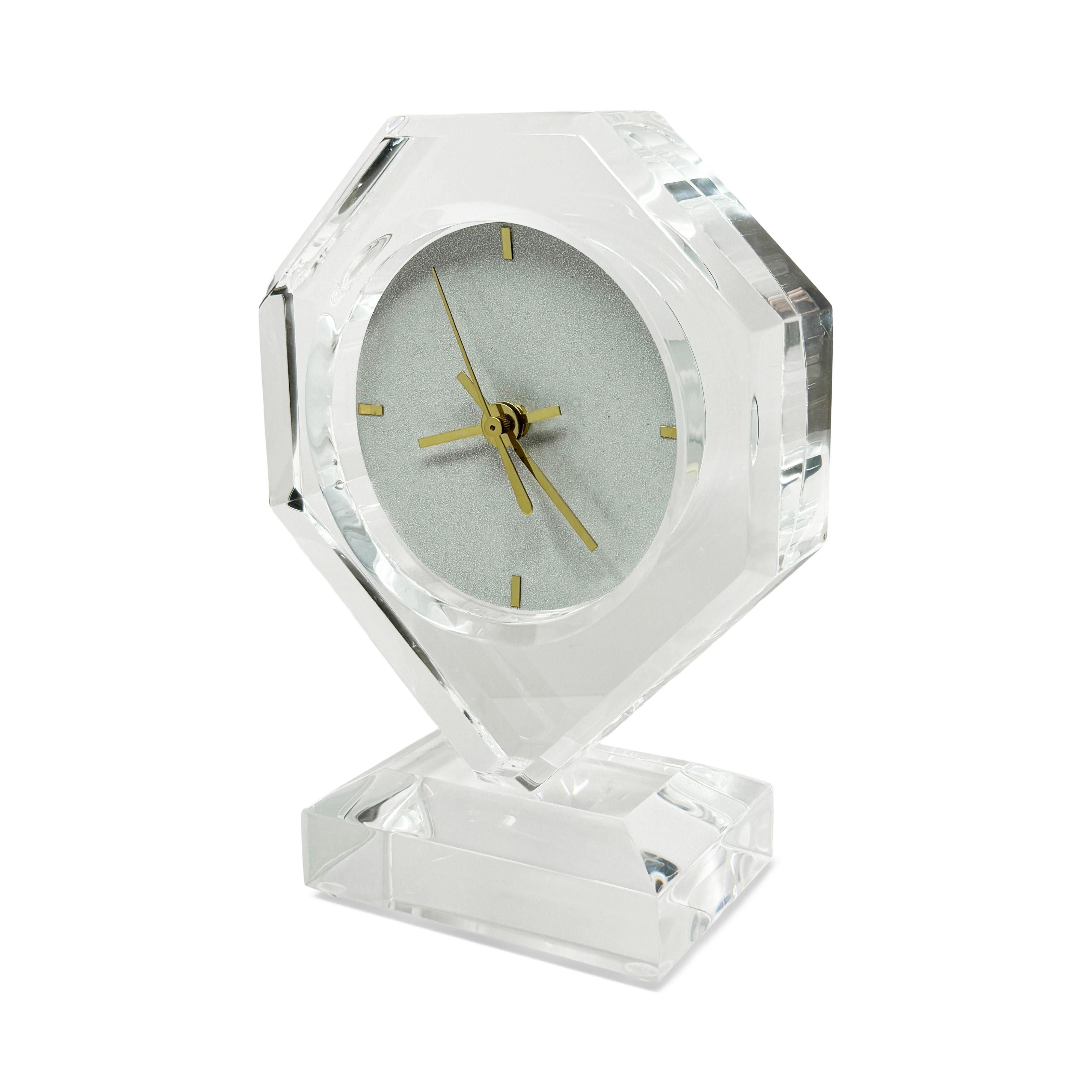 A simple and sophisticated lucite mantle or table clock with a rectangular base, tall diamond shaped body, textured light gray face, brass marks for numbers, and brass hands.  It is sleek with clean lines, simple elegance, and a perfect modern