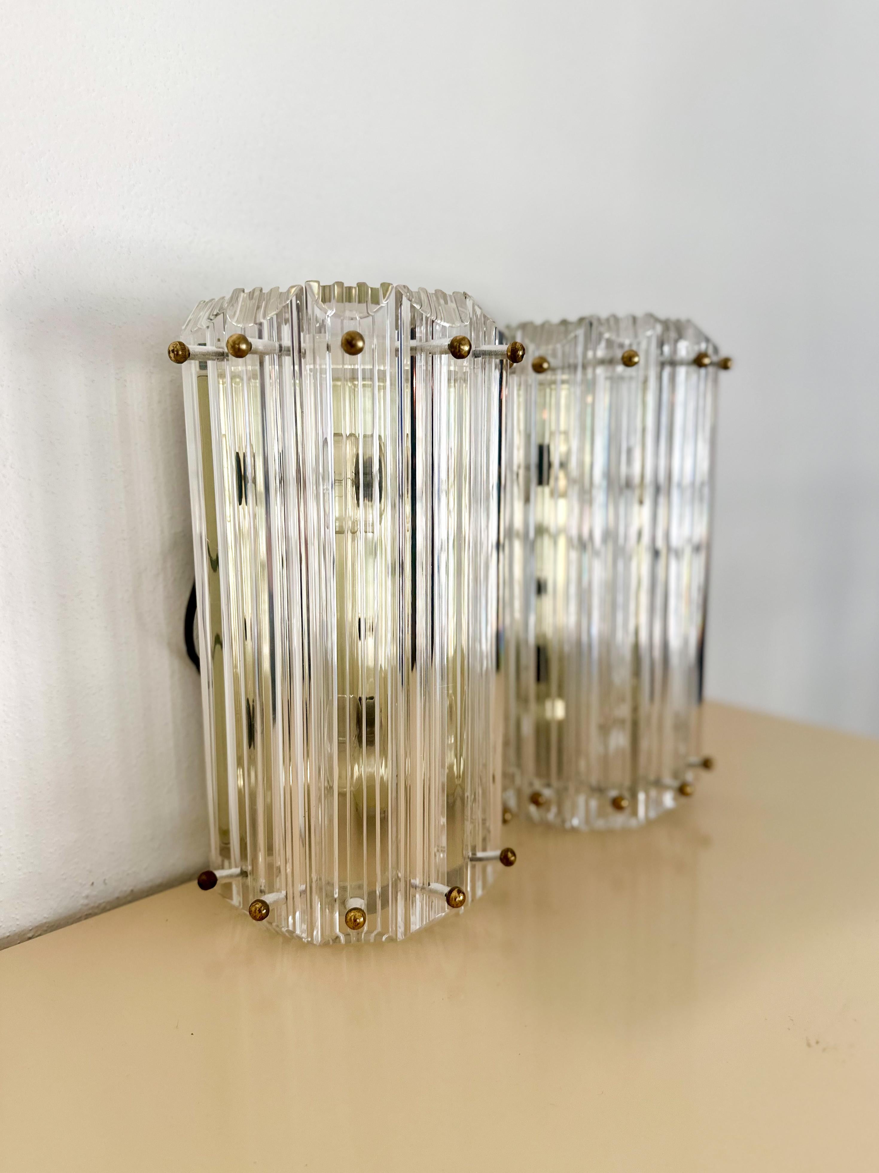 Beautiful pair of Hollywood Regency prismatic lucite wall mounted sconces with brass detail, made by Triarch Industries in Miami, c.1989. Each sconce has 7 lucite panels, held together by brass ball-cap screws, and holds two bulbs. In excellent