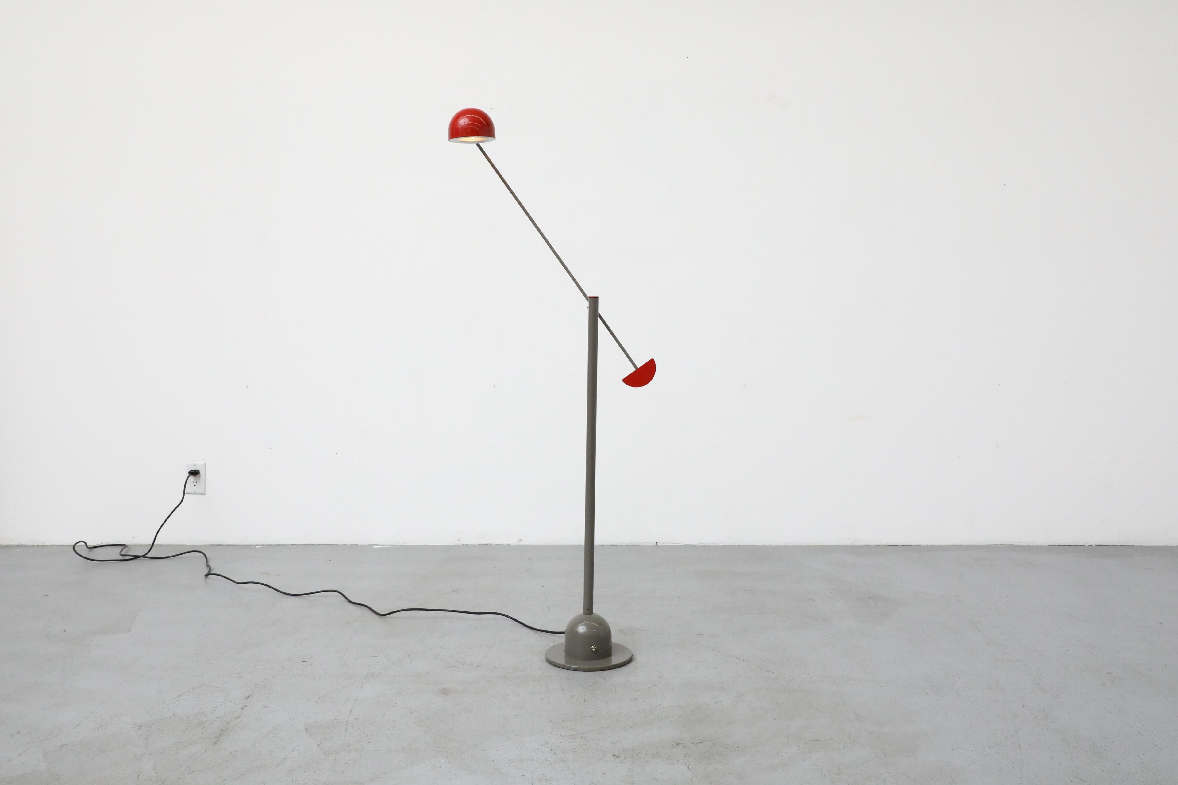A playful floor lamp inspired by Memphis Design from the 1980s, with a gray enameled frame and base, a cantilevered arm, and a geometrically shaped, half-dome red enameled metal shade. It is reminiscent of German lighting manufacturer Honsel