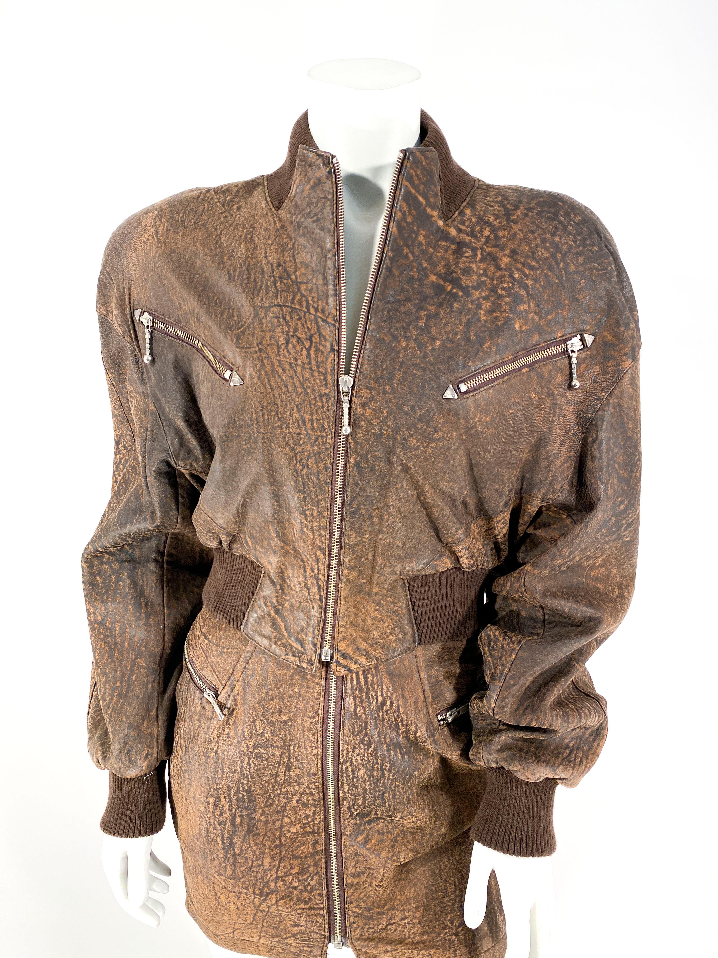 1980s North Beach Leather brown bomber jacket with zip closure and heavily padded shoulders, oversized/dramatic shape, and wide ribbing. The matching skirt has zip pocket and closure. The entire set is made of a soft brown leather with a distressed