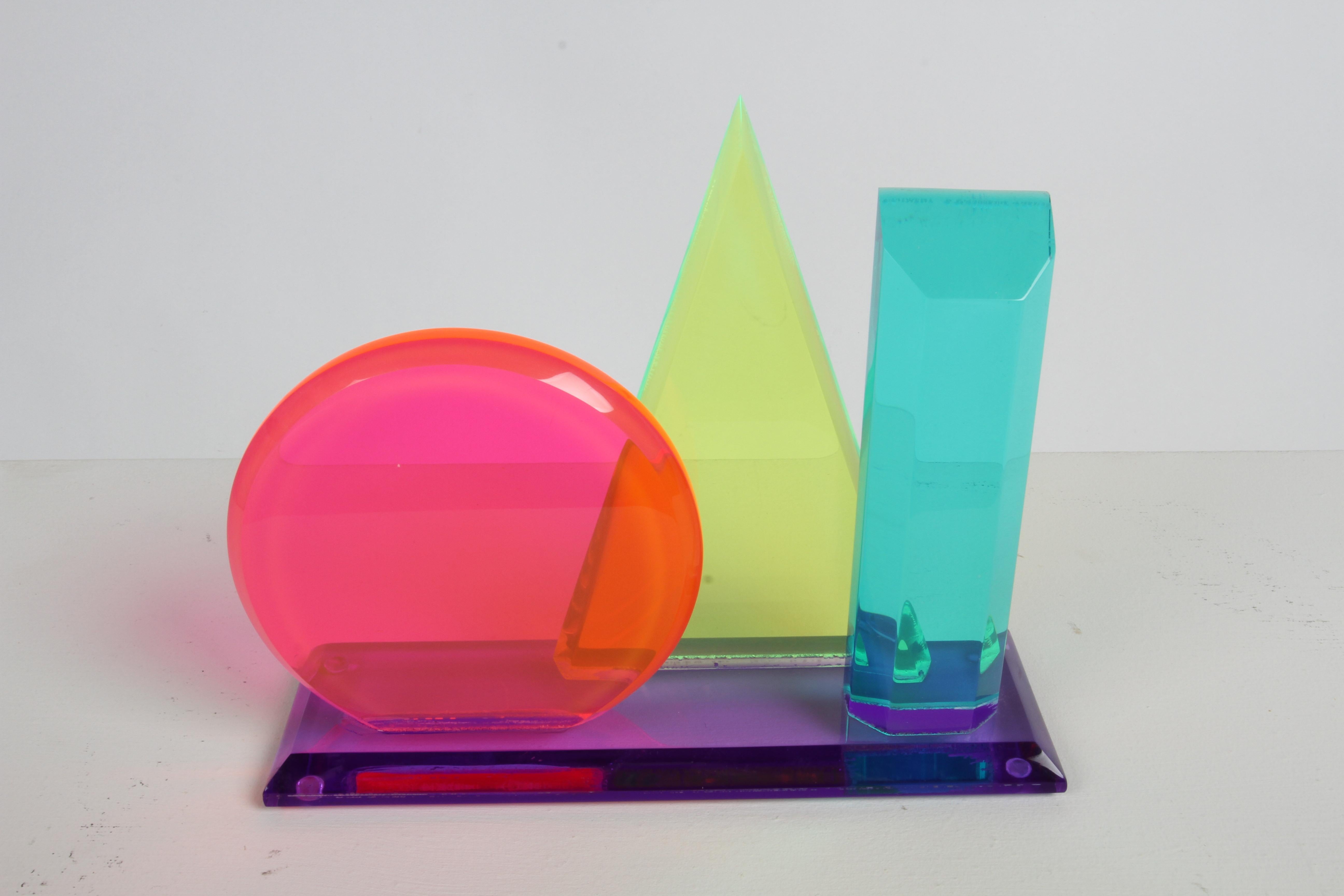 In the style of Vasa Mihich or Shlomi Haziza, 1980s sculpture colored beveled shapes made of acrylic or lucite with pink circle, yellow triangle, teal rectangle on a purple base. This makes a great desk accessory or a sculptural object for a