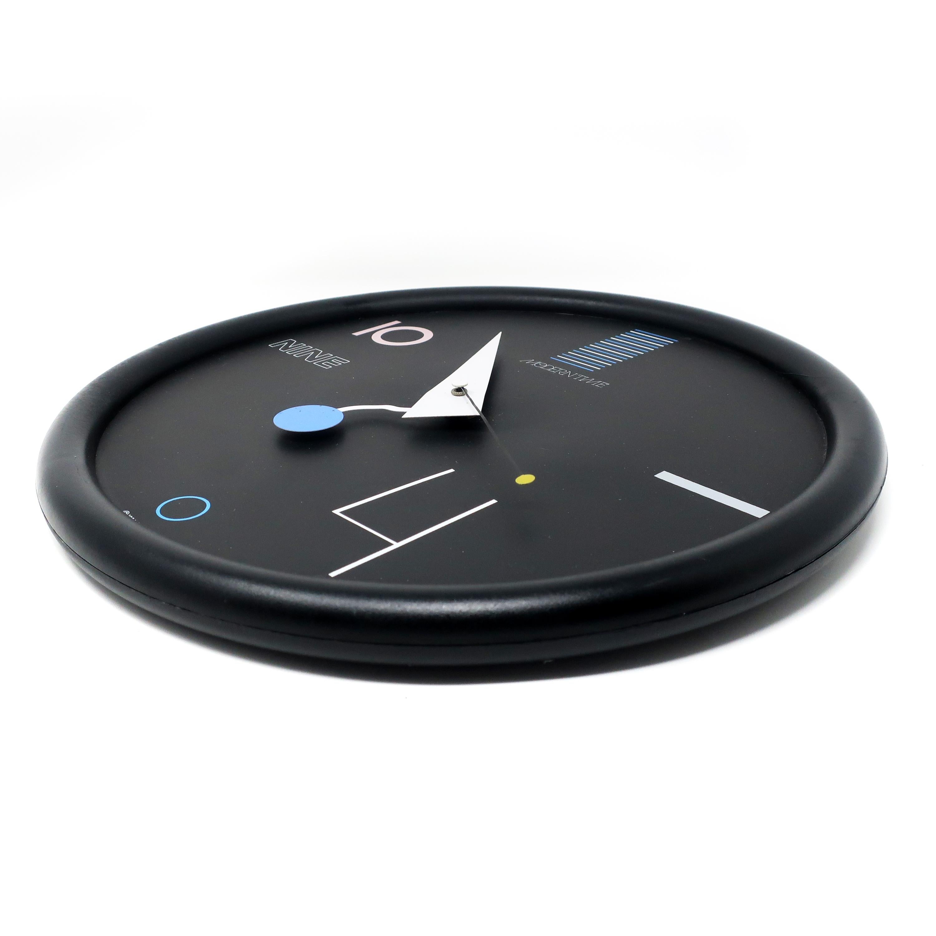 A stunning postmodern wall clock with multi-color accents from Canetti's Moderntime Collection. Black plastic frame, black face, and blue, white, black, and gray accents. It perfectly walks the line between understated & sophisticated and bright &