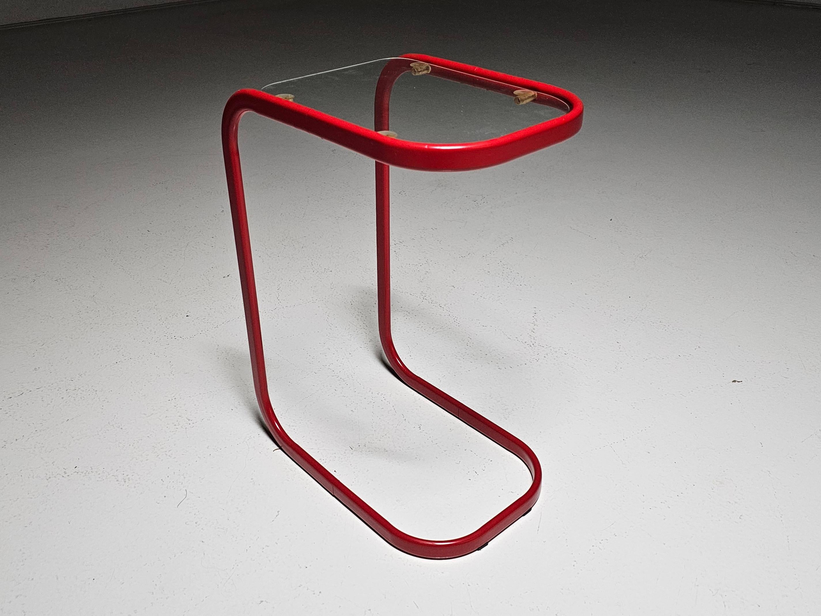 This sleek 1980s side table is designed for effortless convenience. Its minimalist tubular frame easily slides over the arm of a sofa or chair, creating an instant surface for your drink, book, or laptop. The red metal frame and glass top give it a