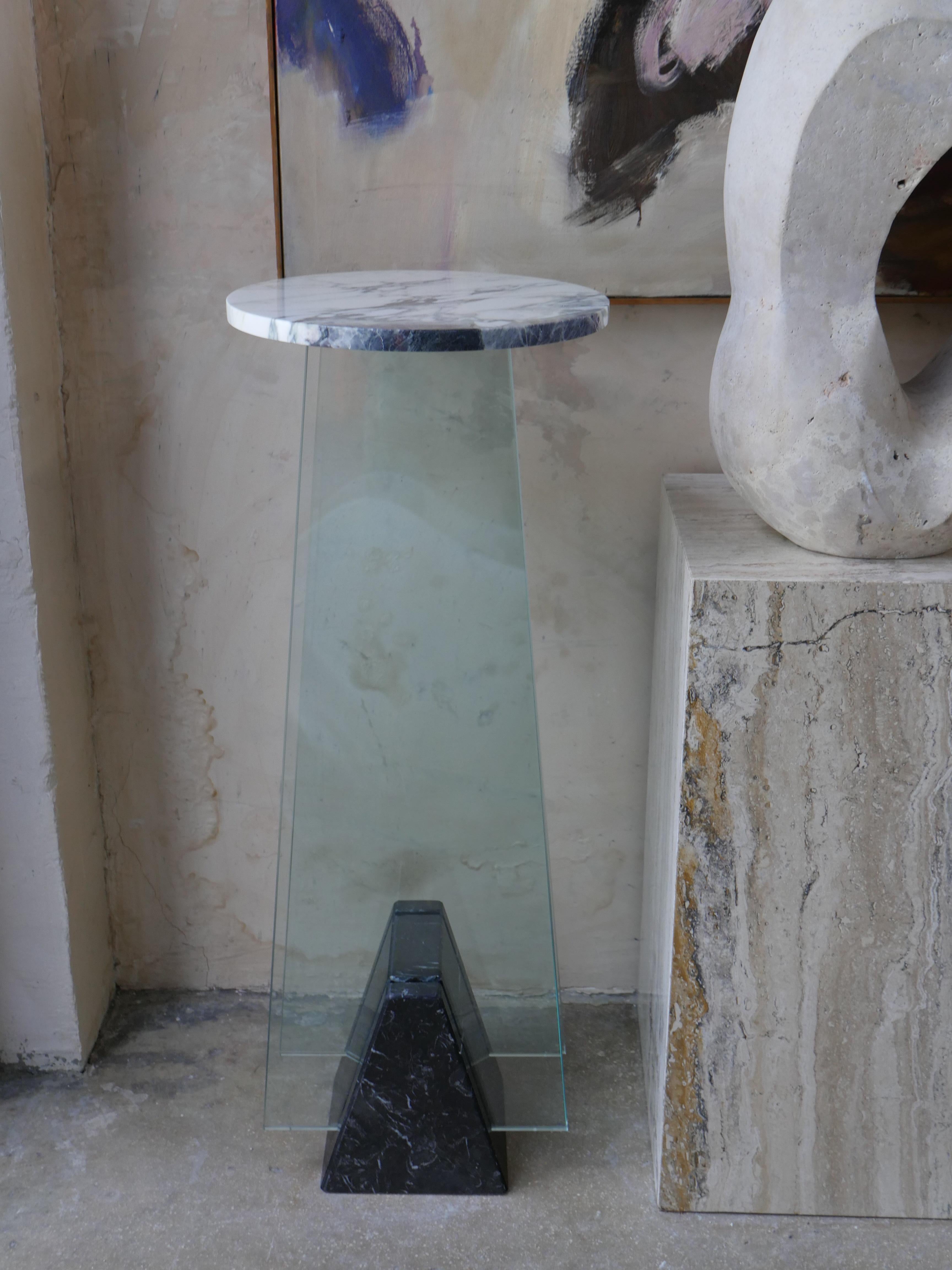 A striking 1980s postmodern pedestal that blends bold materials and geometric precision. Featuring a Breccia Capraia marble top with rich veining, a sleek Nero Marquina marble base, and a glass foundation that adds a dynamic, architectural edge.